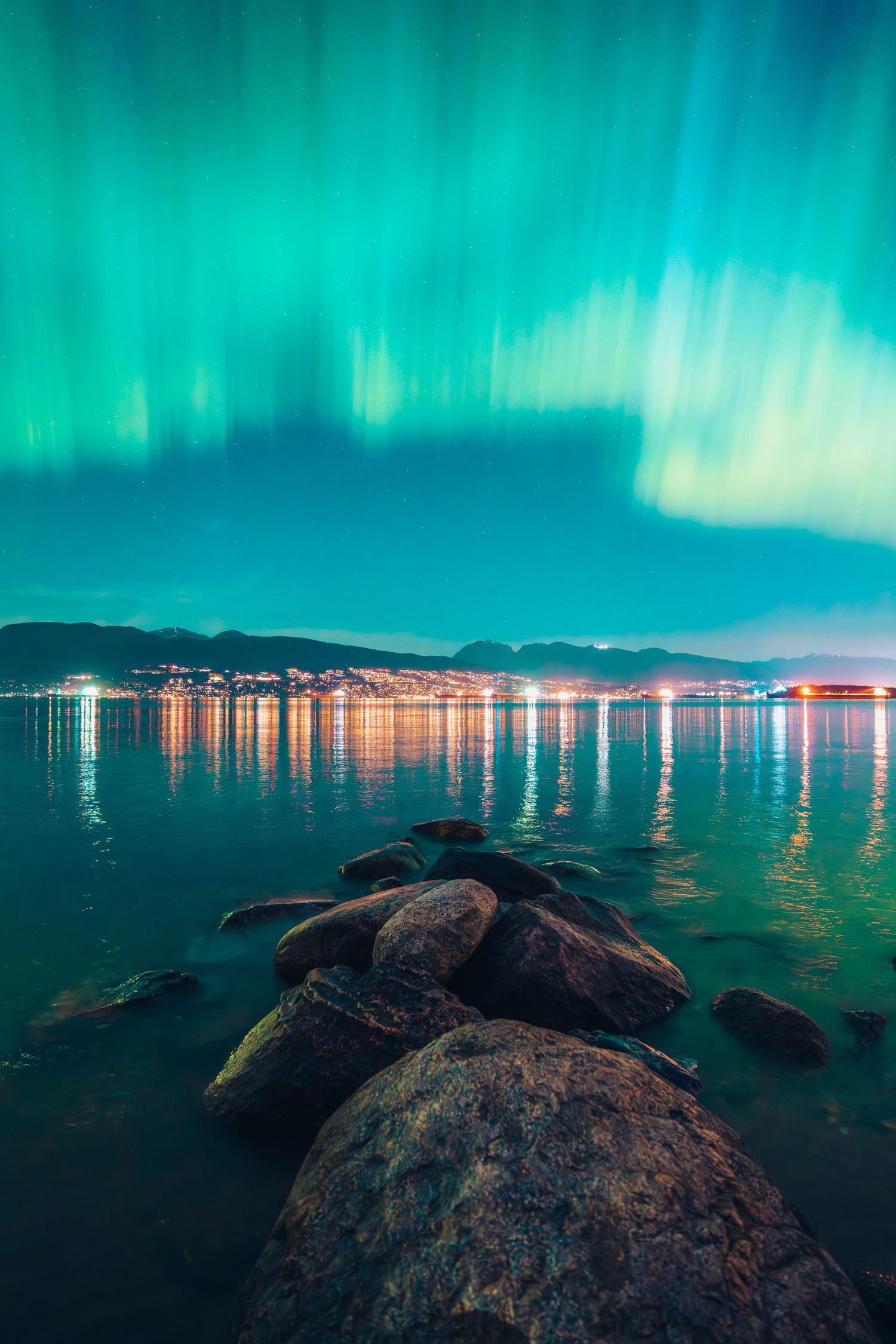 Vivid teal aurora borealis reflects over calm water with city lights glowing in the distance.