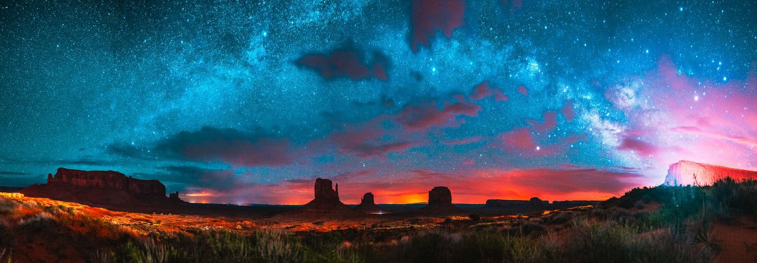 Panoramic night sky with Milky Way over Monument Valley's iconic buttes, vibrant red and blue hues at twilight.