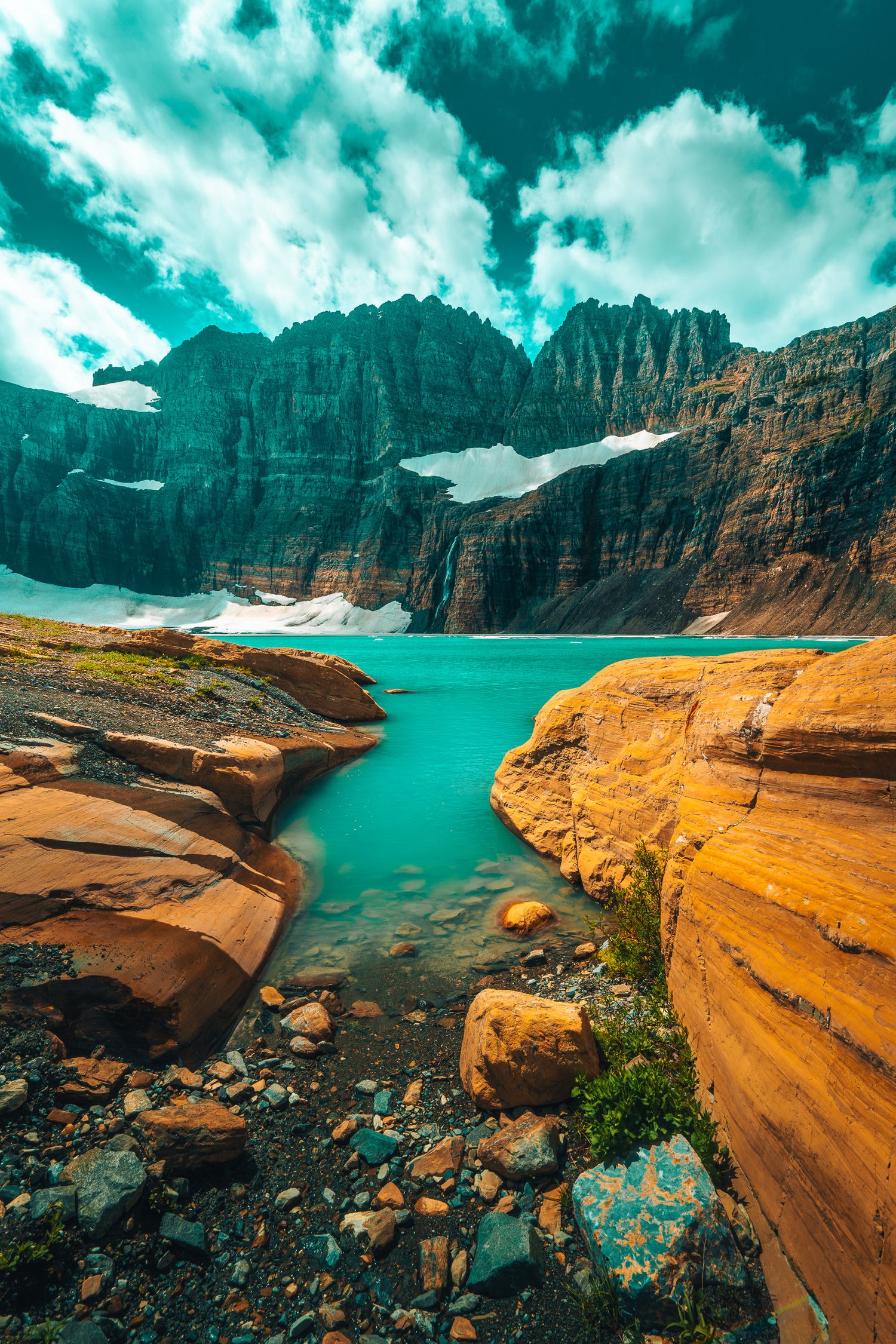 Turquoise glacial lake surrounded by rugged orange rocks and snow-capped mountain peaks under dramatic cloudy skies.