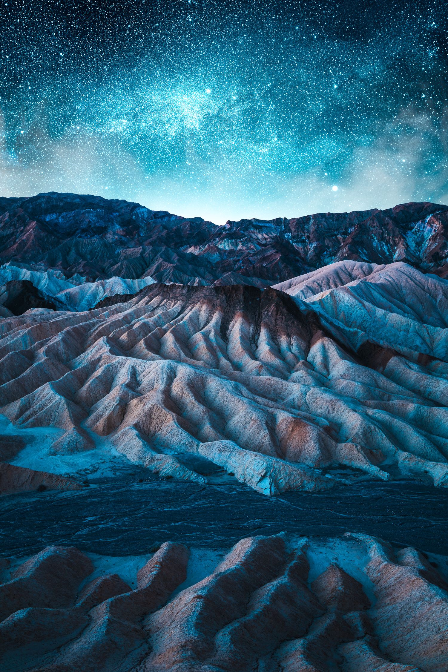 Dramatic eroded badlands rock formations at Zabriskie Point glow under a brilliant blue starry night sky.