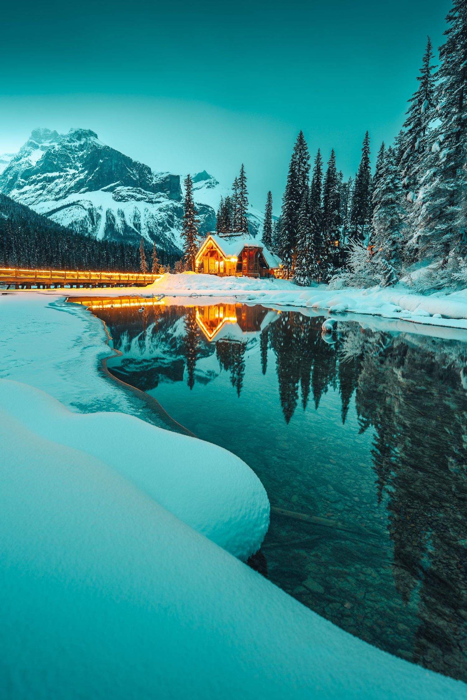 Glowing lodge reflects in a partially frozen lake surrounded by snow-covered pines and mountains.