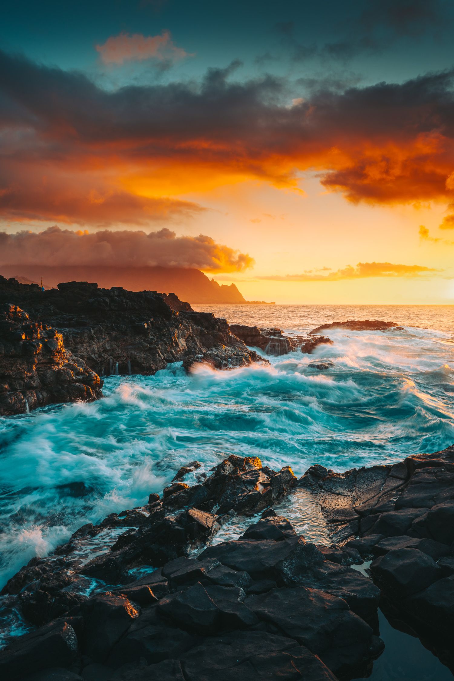 Powerful turquoise waves crash against volcanic rocks during a vivid orange sunset on a rugged coastline.