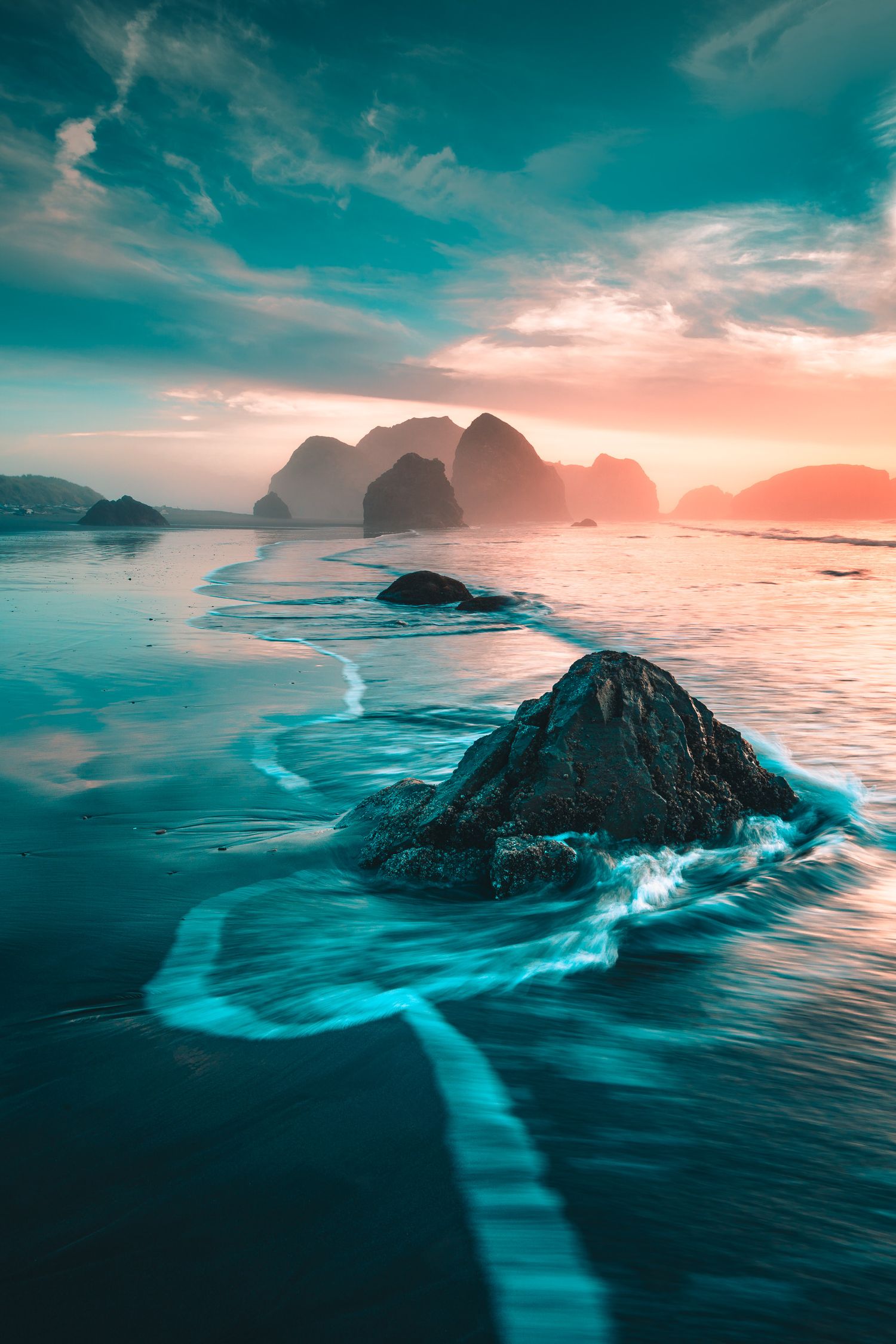 Turquoise ocean waves around rocky outcrops at sunset with misty island silhouettes.