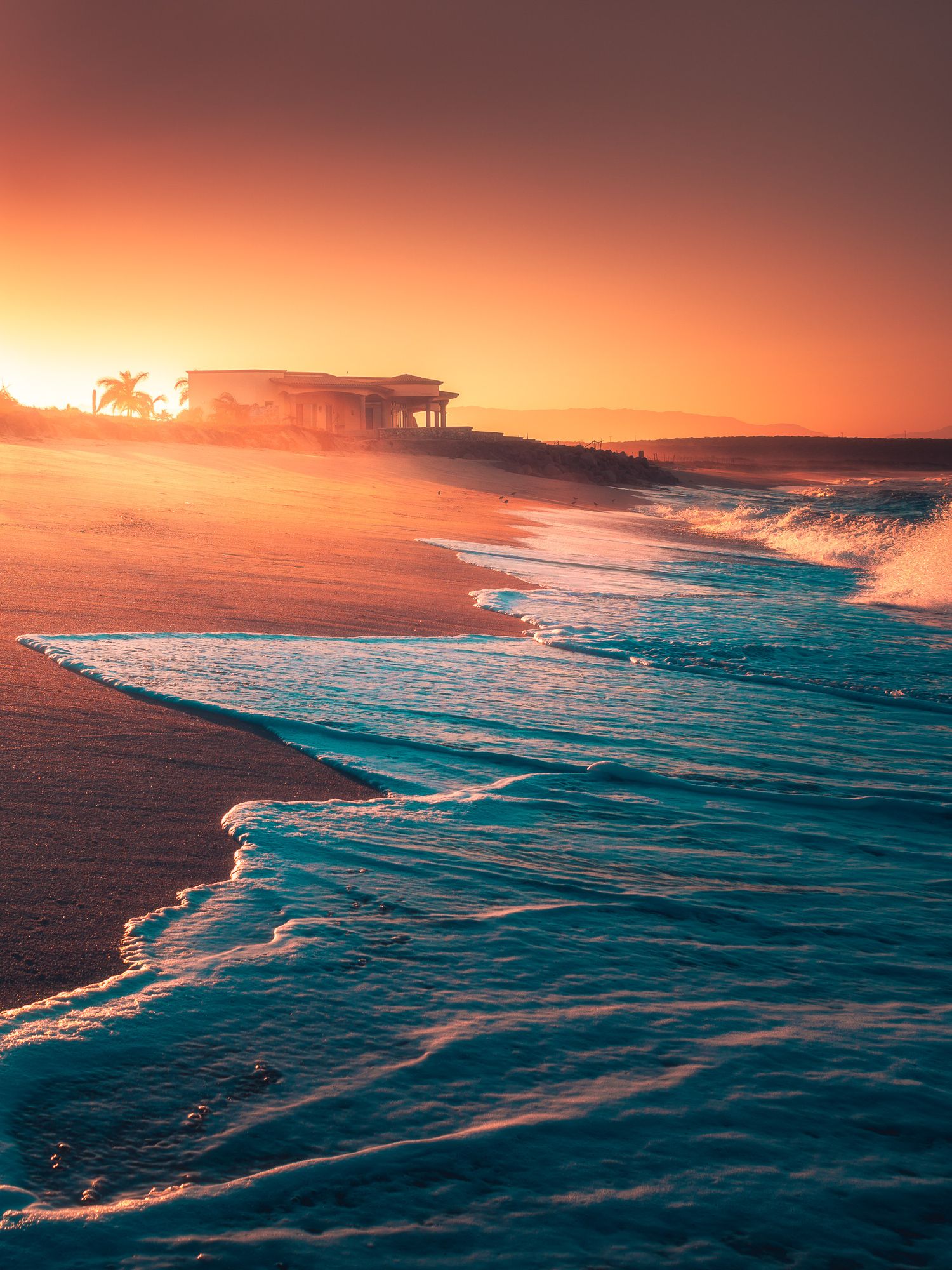 Stunning tropical beach sunset with turquoise waves washing ashore and a silhouetted building glowing in orange light.
