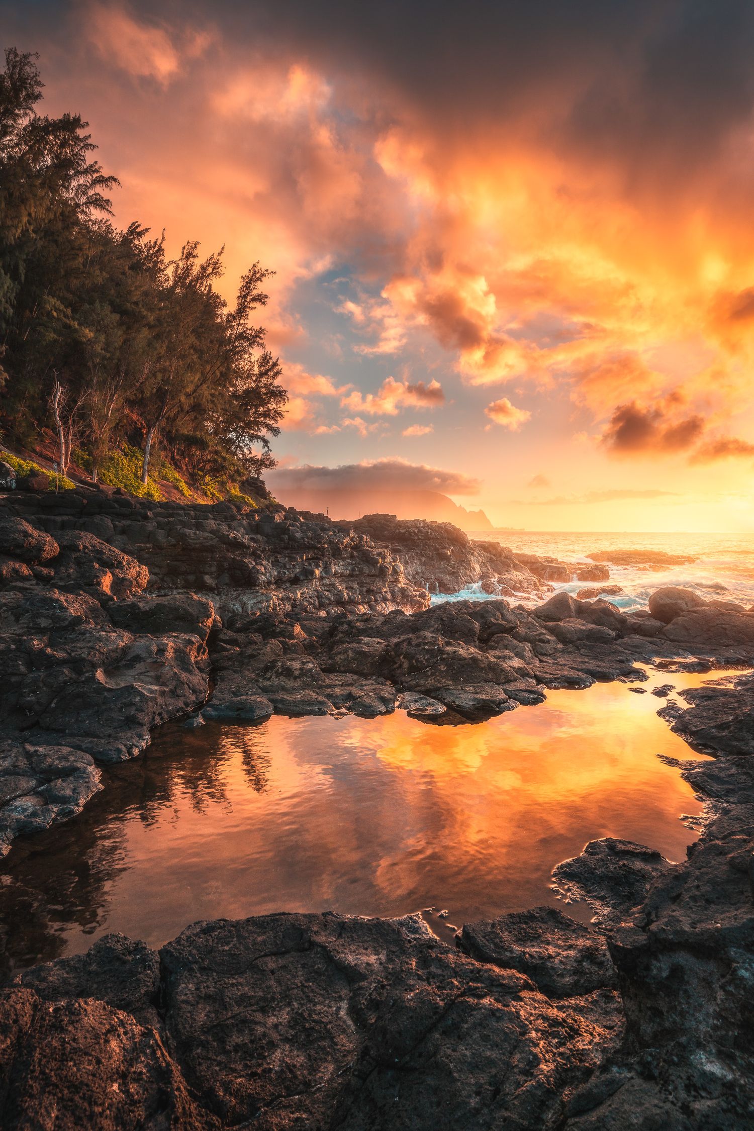 Dramatic sunset over rocky Hawaiian coastline with fiery orange clouds reflected in tidal pools, lush trees silhouetted on left.