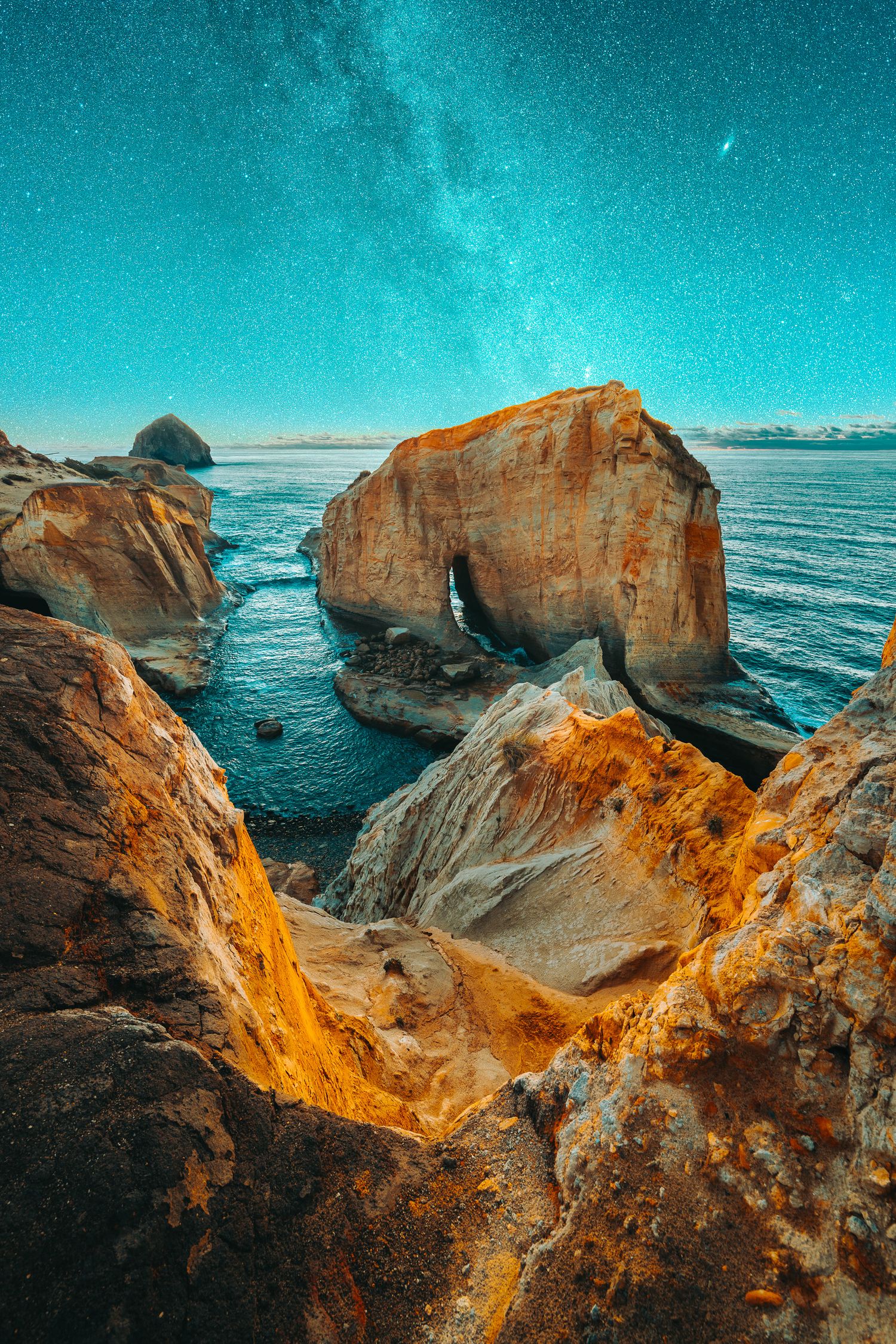 Natural rock arch formation on a coastal cliff glows under a starry Milky Way night sky.