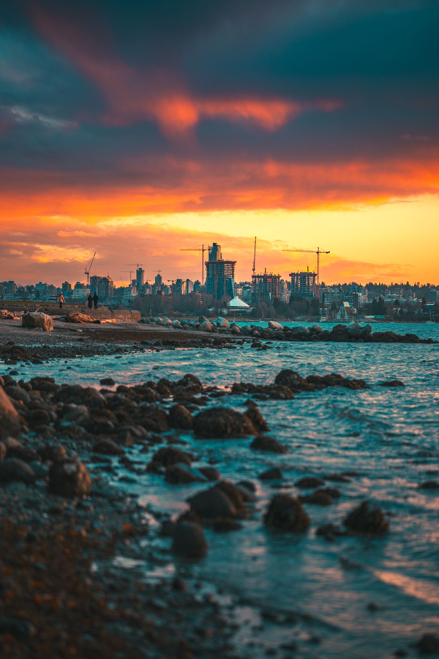 Dramatic sunset over a rocky coastline with construction cranes and city silhouettes under vivid orange and red clouds.