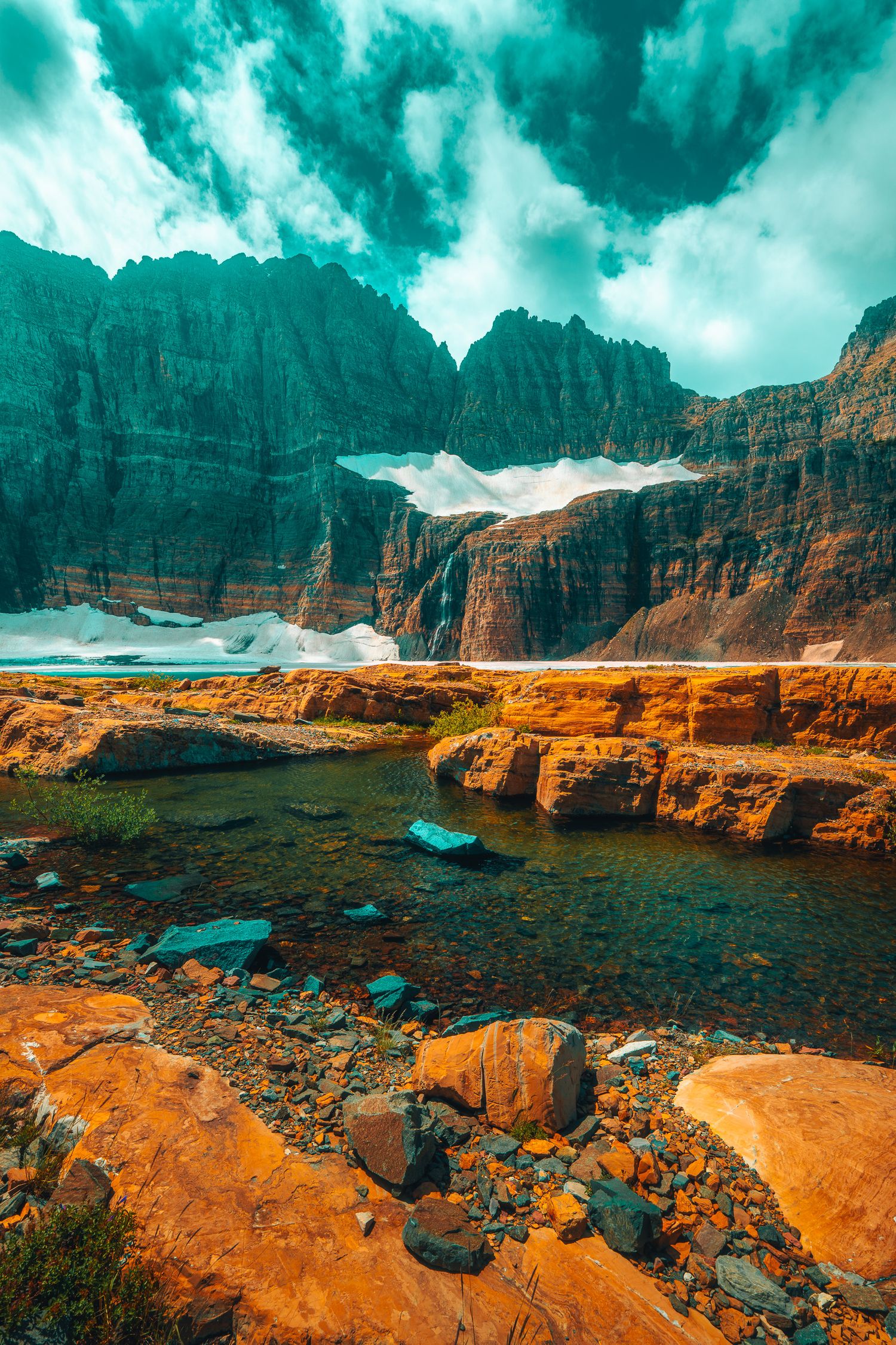 Rocky shoreline with orange boulders and shallow stream beneath towering glaciated cliffs under dramatic teal clouds.