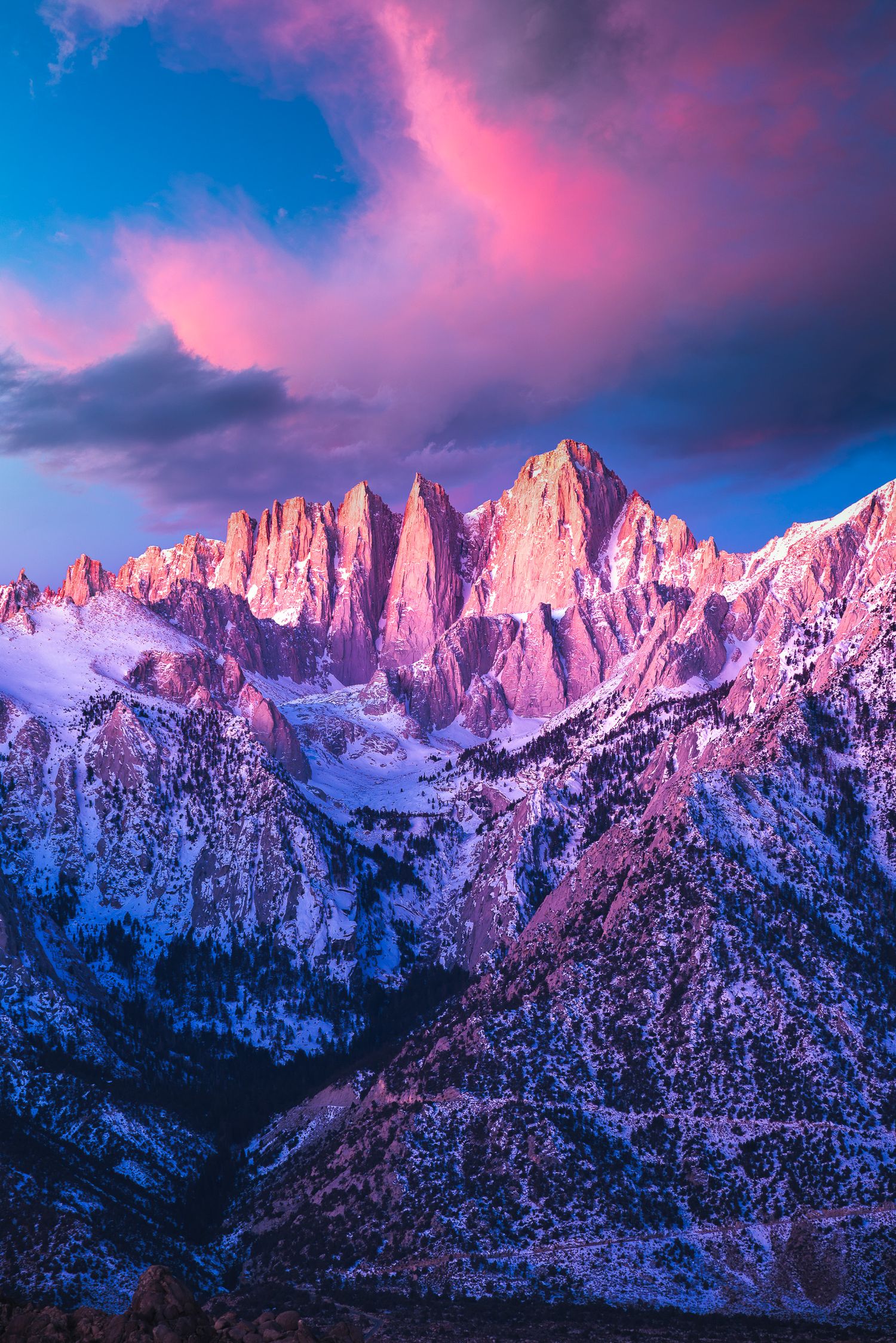 Snow-dusted jagged mountain peaks bathed in pink and purple alpenglow at sunset under a dramatic cloudy sky.