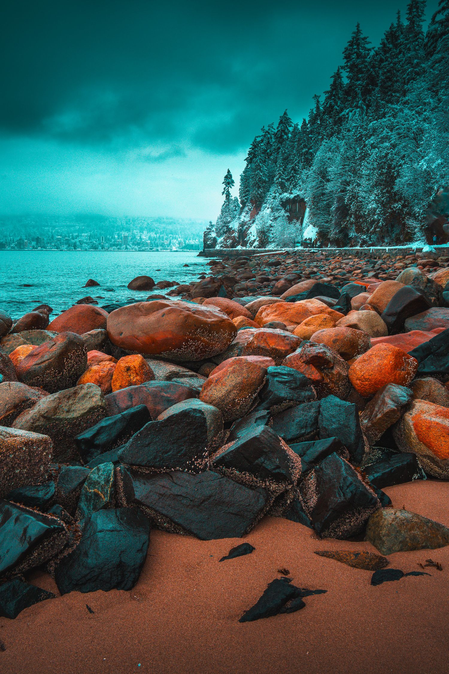 Colorful orange and teal boulders on a sandy shoreline with frost-covered pine trees under a stormy sky.