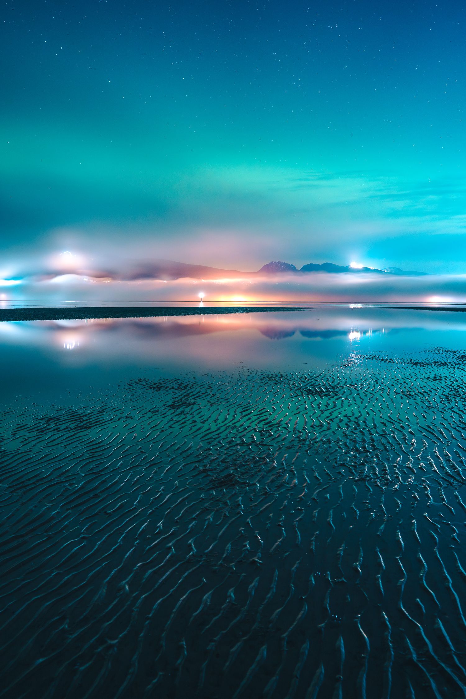Northern lights cast teal and orange hues over a perfectly still reflective beach with rippled sand at low tide.