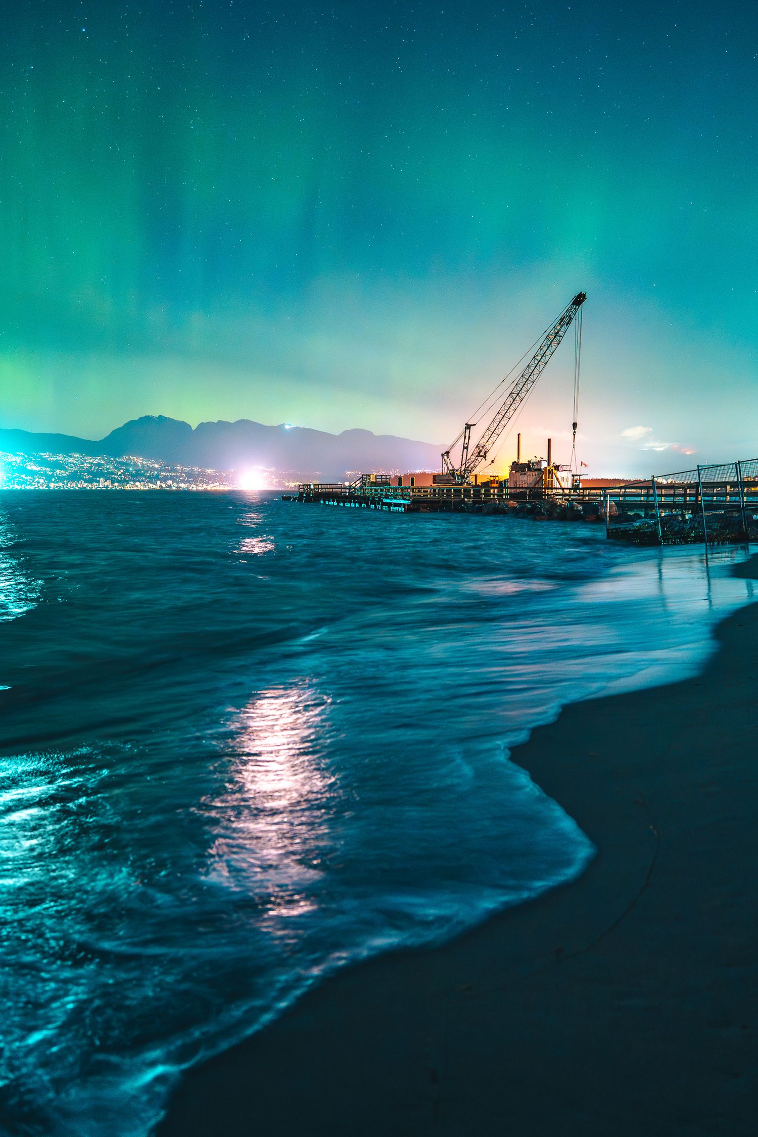 Northern lights illuminate a coastal pier with a crane under a teal aurora borealis sky.