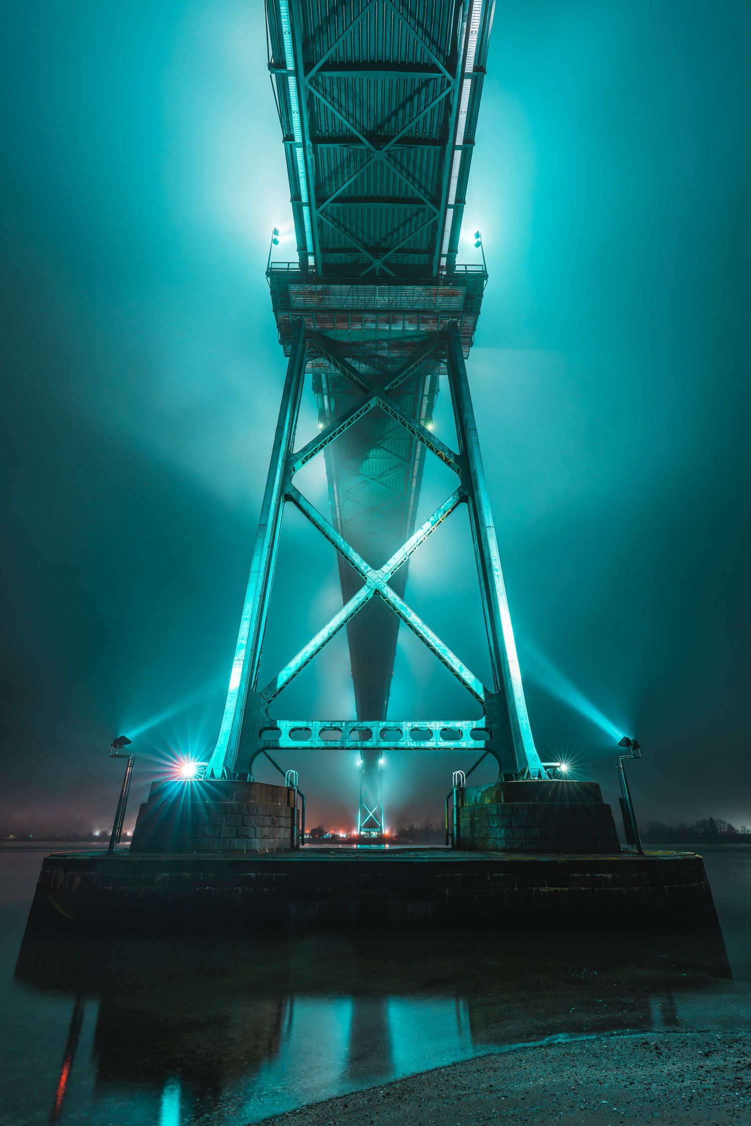 Low-angle view of a massive bridge support tower glowing teal through thick fog above a misty harbor.