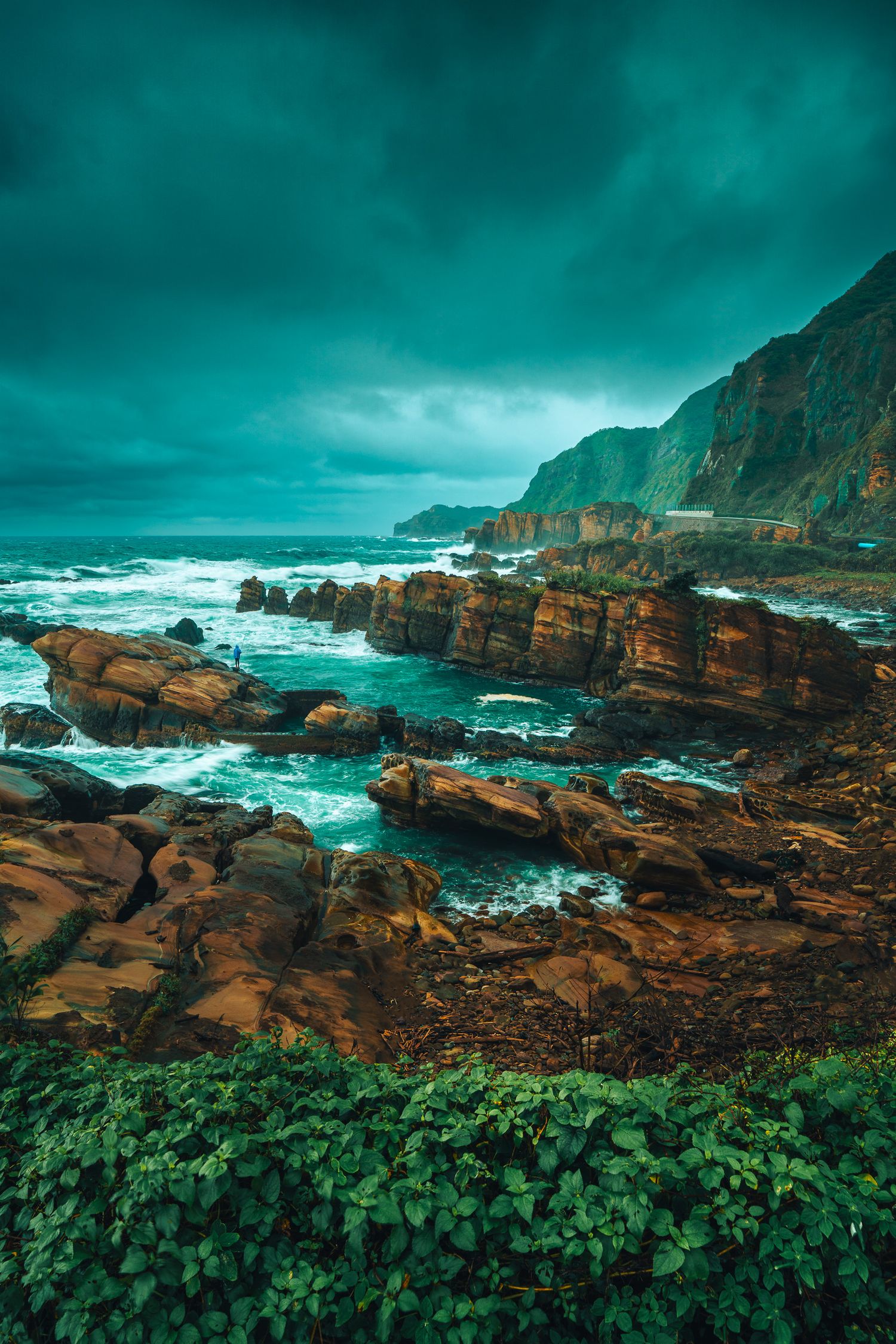 Rugged coastal cliffs with brown rocks and crashing waves under a stormy teal sky.
