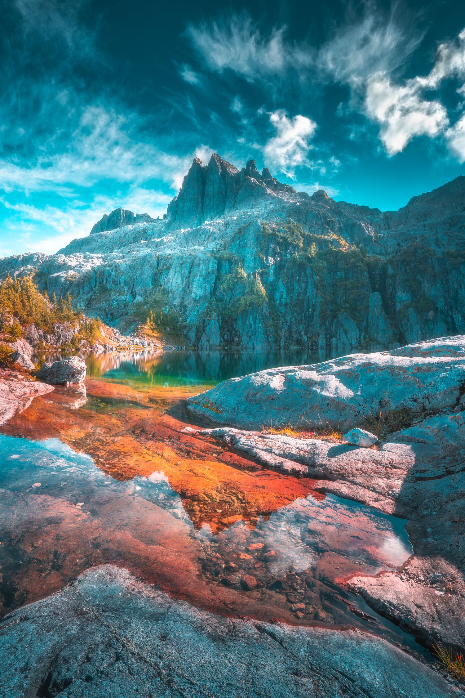 Vivid alpine lake surrounded by colorful rocky terrain and dramatic teal-toned mountain peaks under a cloudy sky.