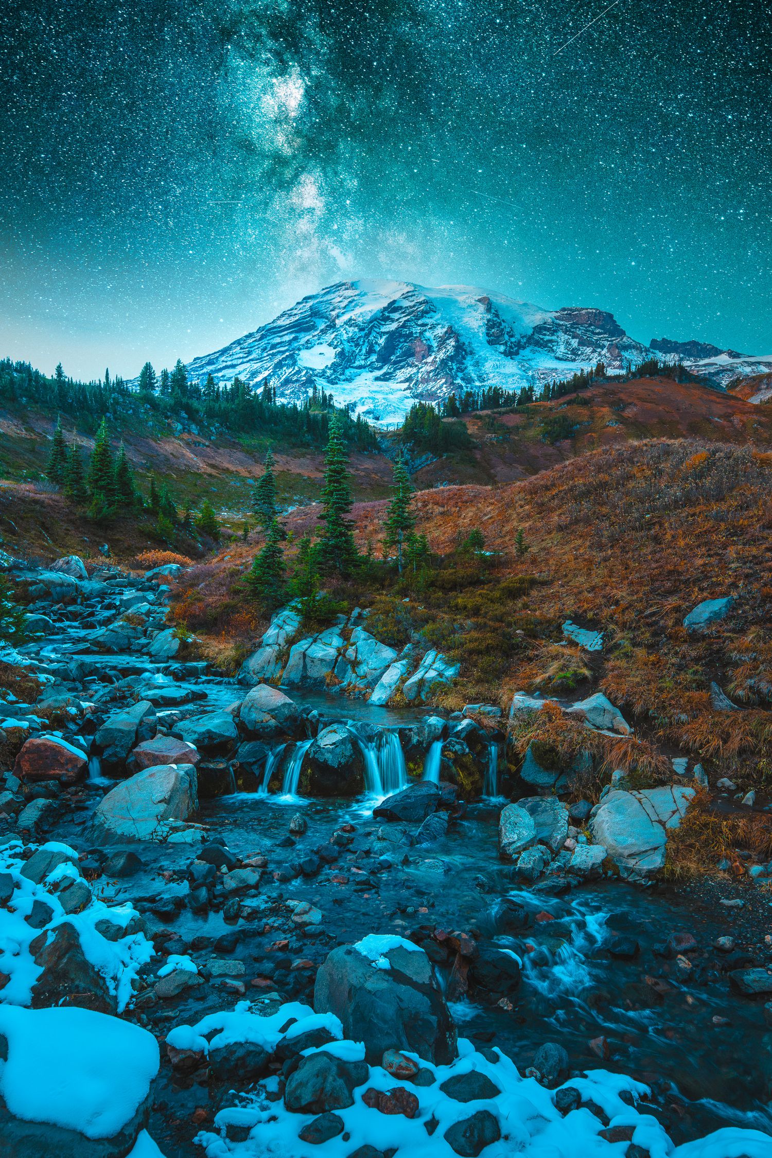 Snow-capped Mount Rainier rises above a rocky stream beneath a brilliant Milky Way starry sky.