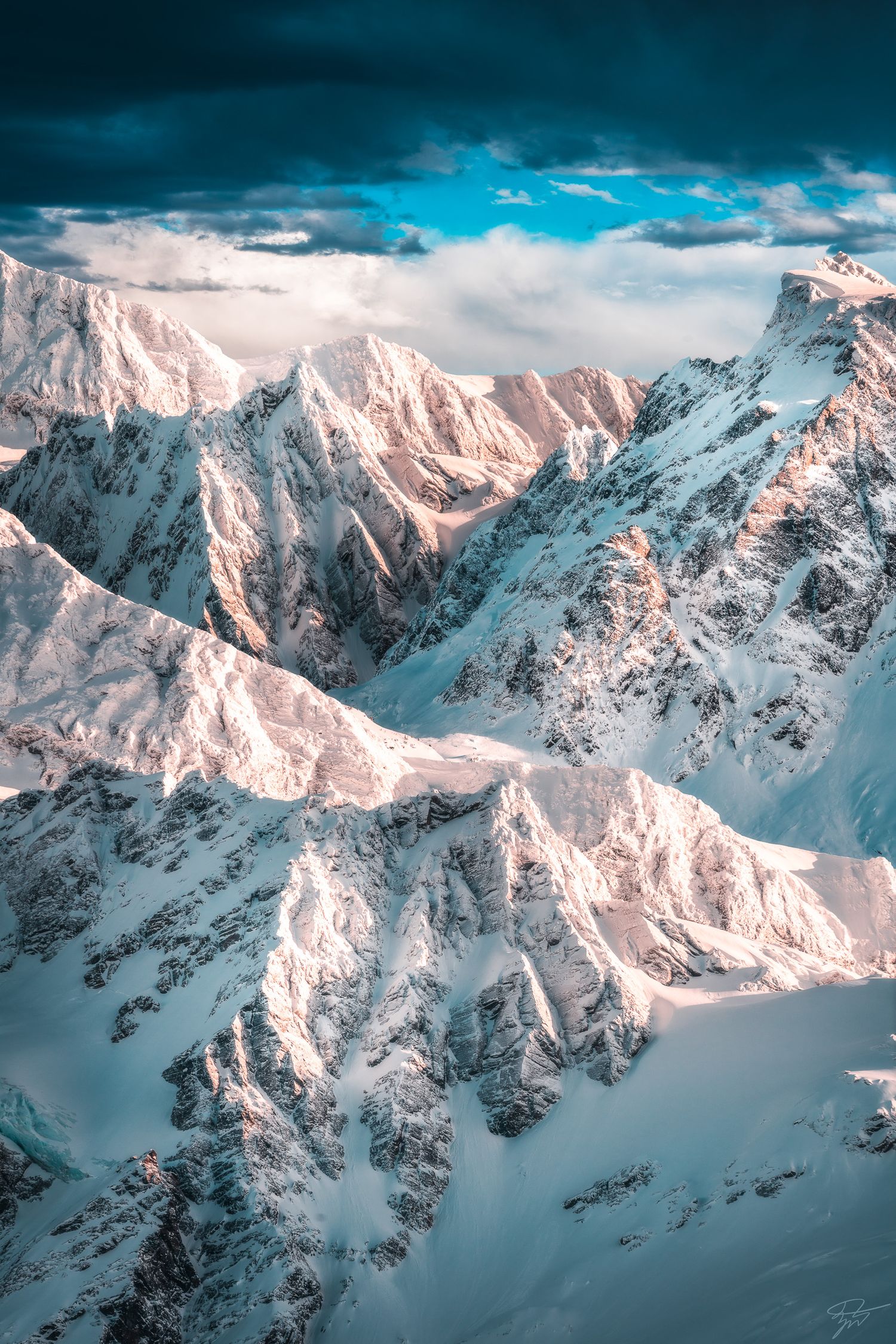 Aerial view of dramatic snow-covered mountain peaks bathed in pink alpenglow under a stormy sky.