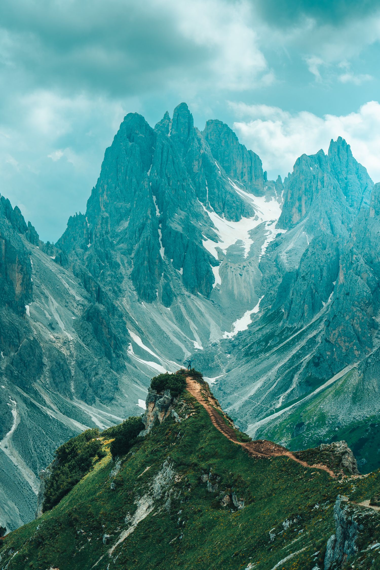 Dramatic Dolomite mountain peaks with snow patches and lush green hillside under a cloudy turquoise sky.