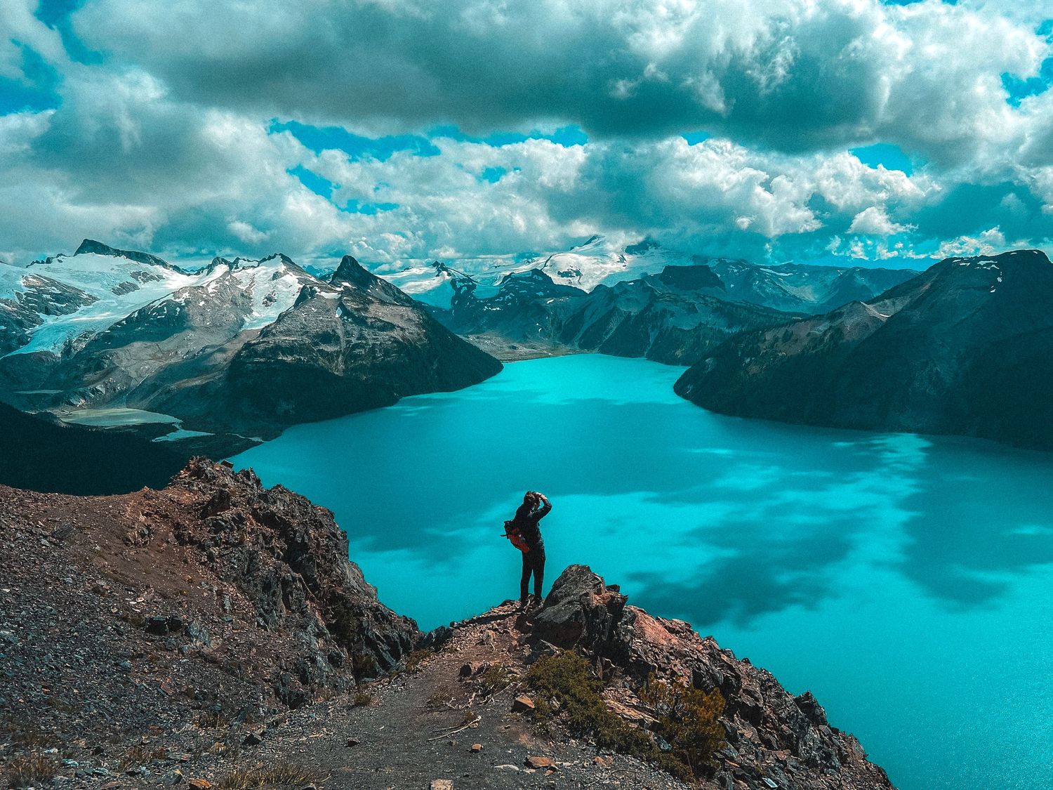 Lone hiker on rugged cliff edge gazing over stunning turquoise Garibaldi Lake and snowy mountains.