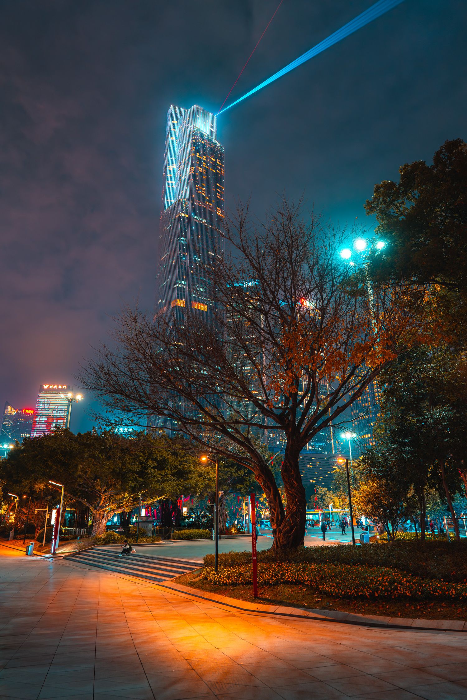 A towering skyscraper with blue laser beams pierces the night sky above an urban park with autumn trees.