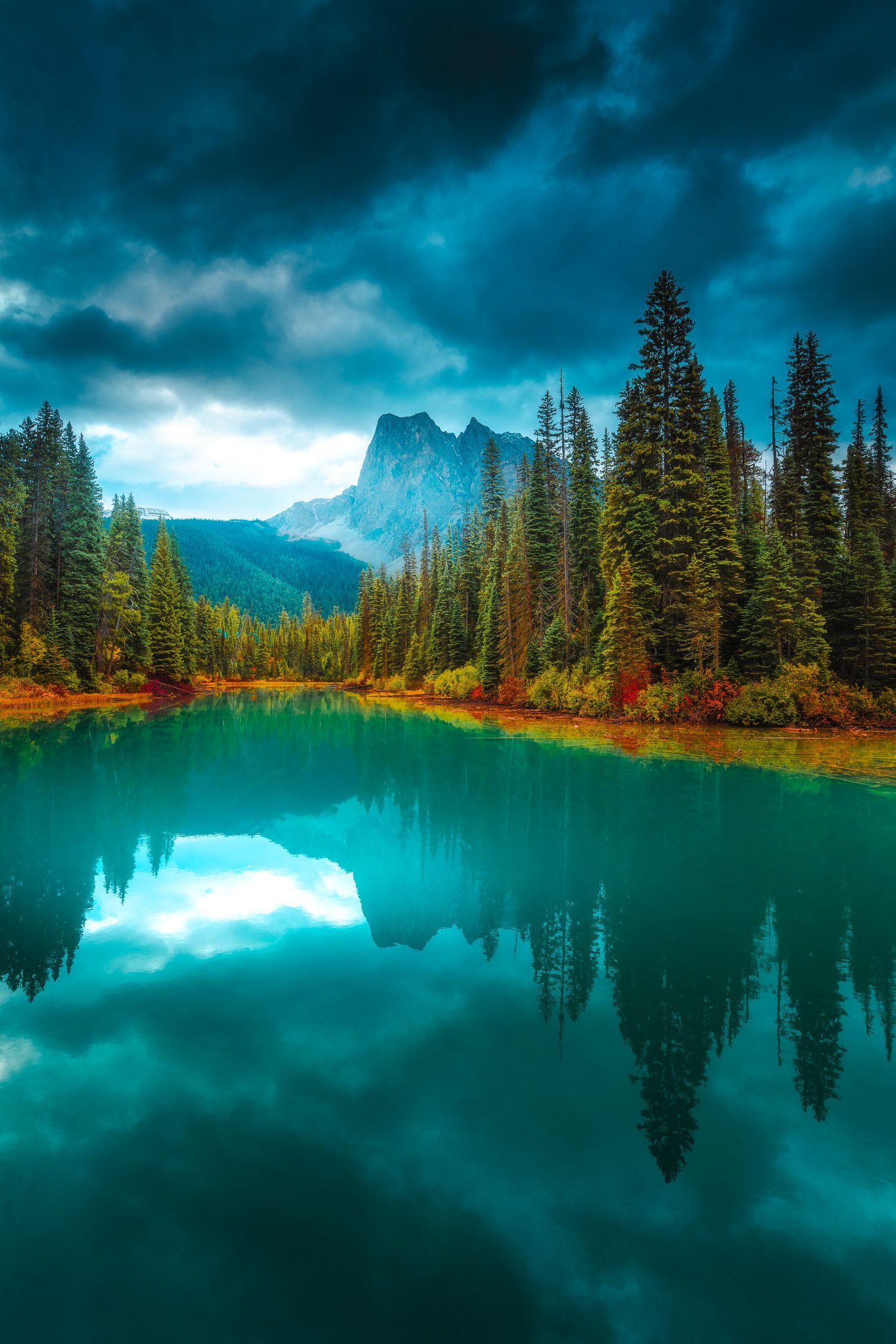 Serene teal mountain lake reflecting pine trees and dramatic storm clouds with a distant rocky peak.
