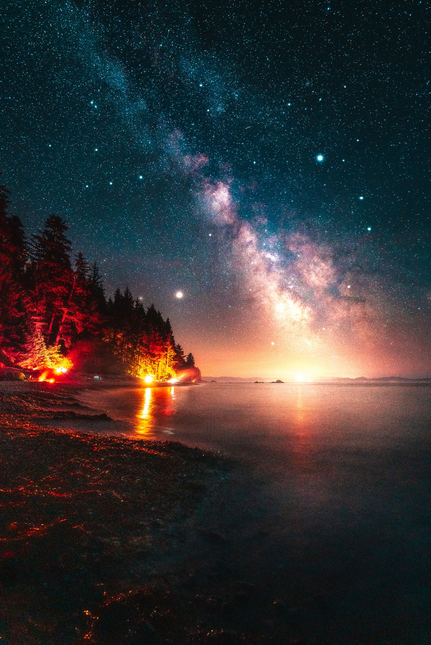 Milky Way galaxy arches over a tranquil coastal forest with a glowing campfire reflecting on the still water below.