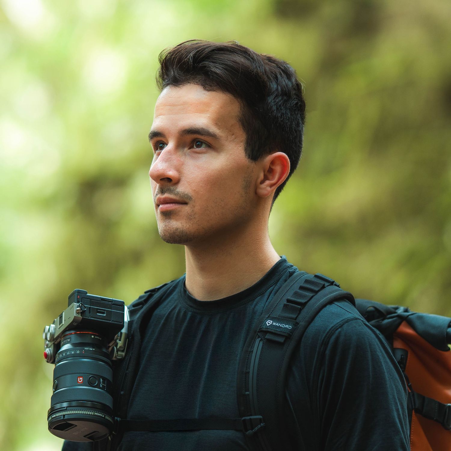 Young male photographer carrying a camera and orange backpack gazing thoughtfully in a green forest.