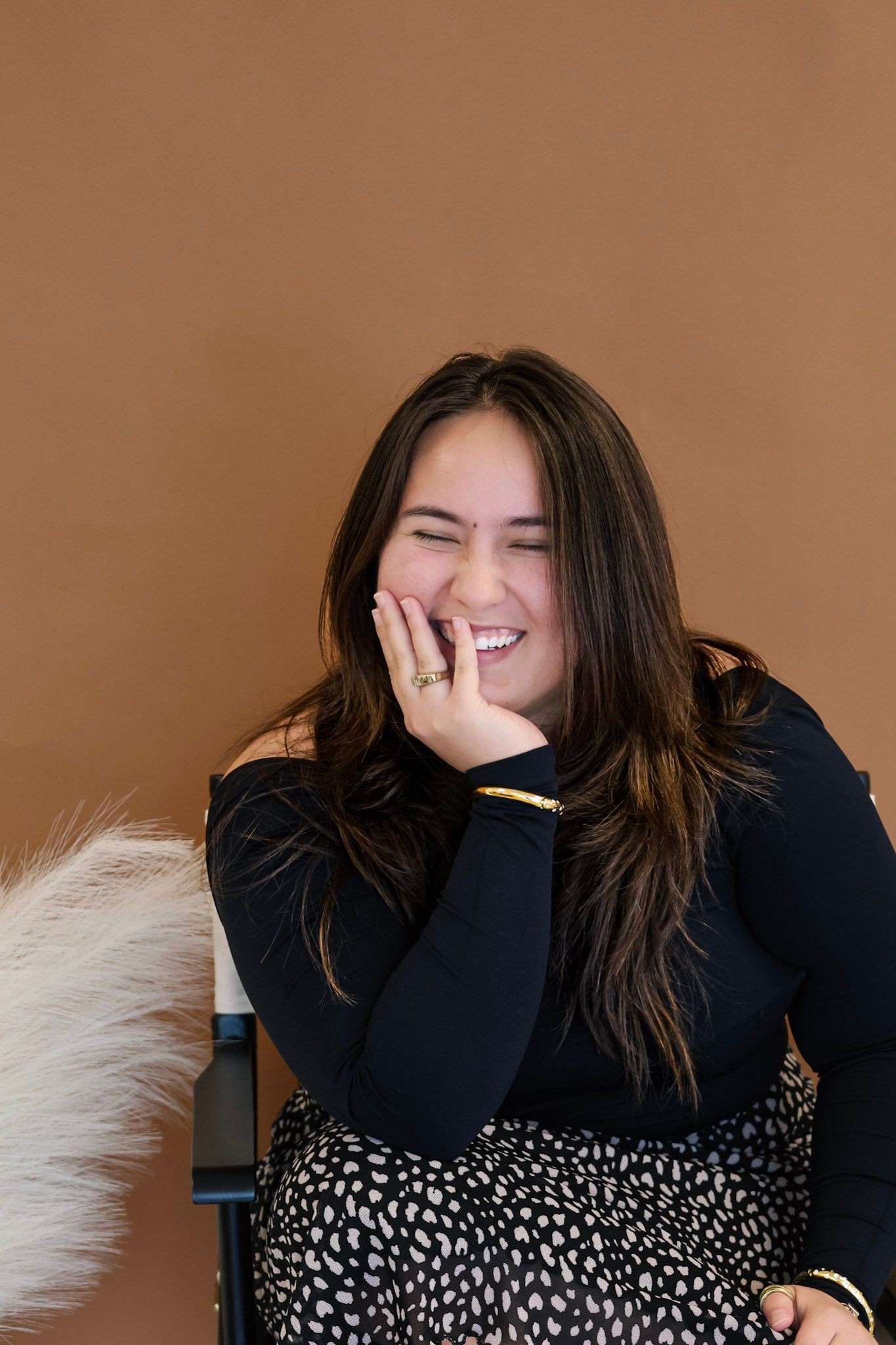 Laughing brunette woman in black top and leopard skirt, resting hand on cheek, seated against warm brown background.