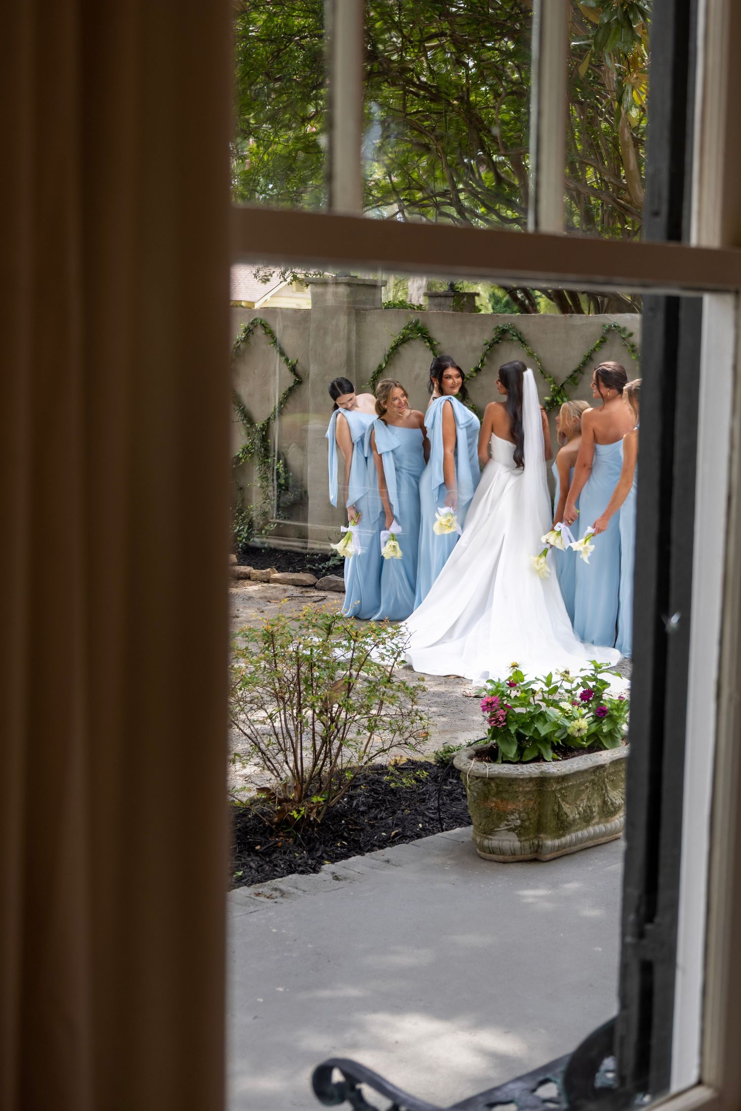 Bride in white gown with bridesmaids in blue dresses posing outdoors, viewed through an open door.