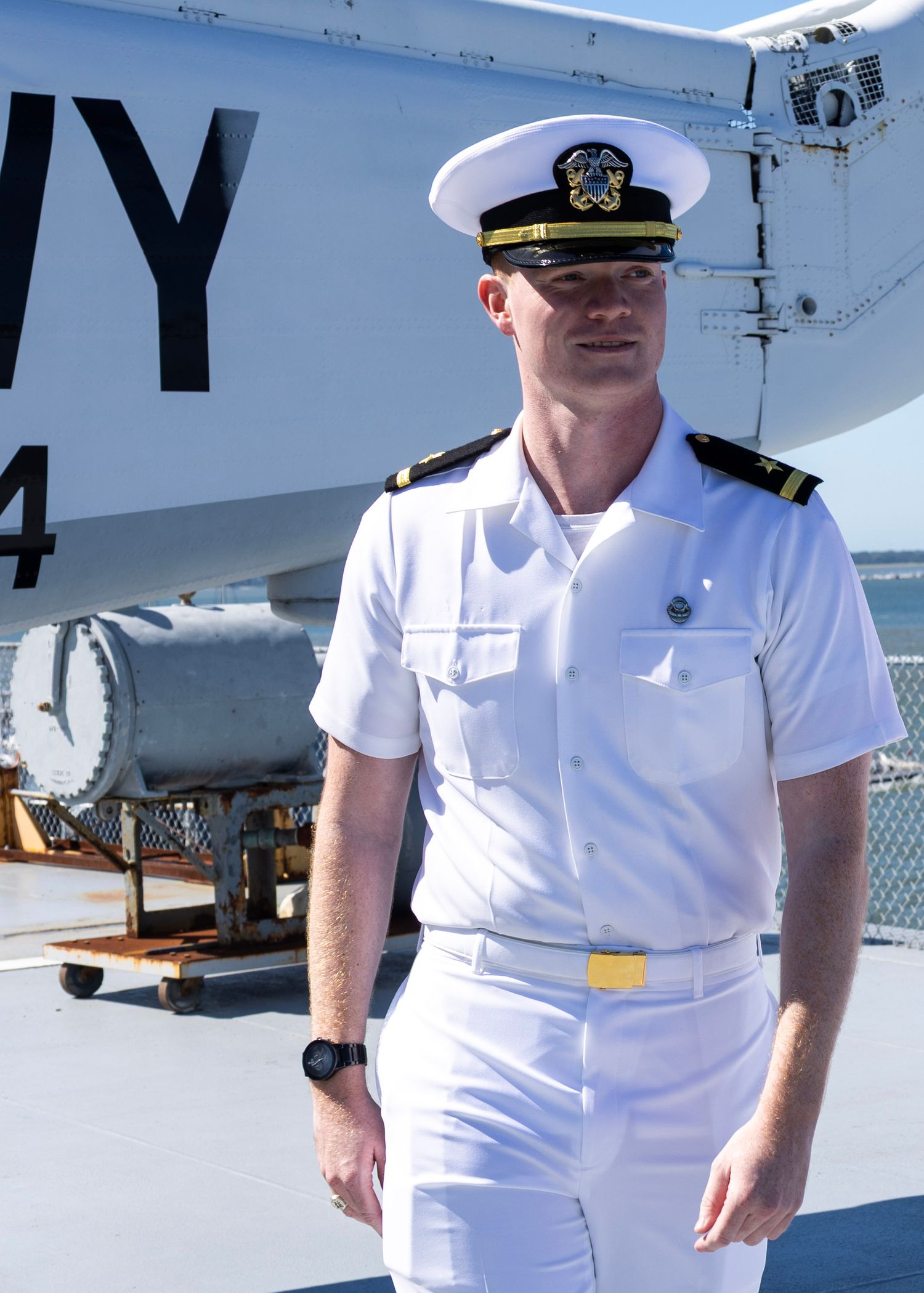 Navy officer in white dress uniform standing on ship deck under clear blue sky.