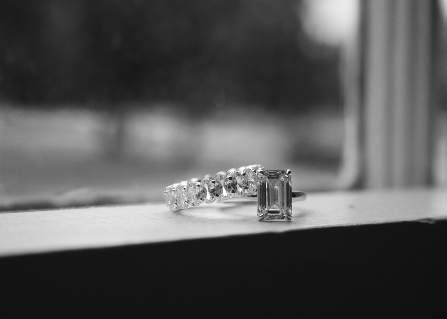 Black and white photo of an emerald cut diamond engagement ring with diamond band resting on a window sill.