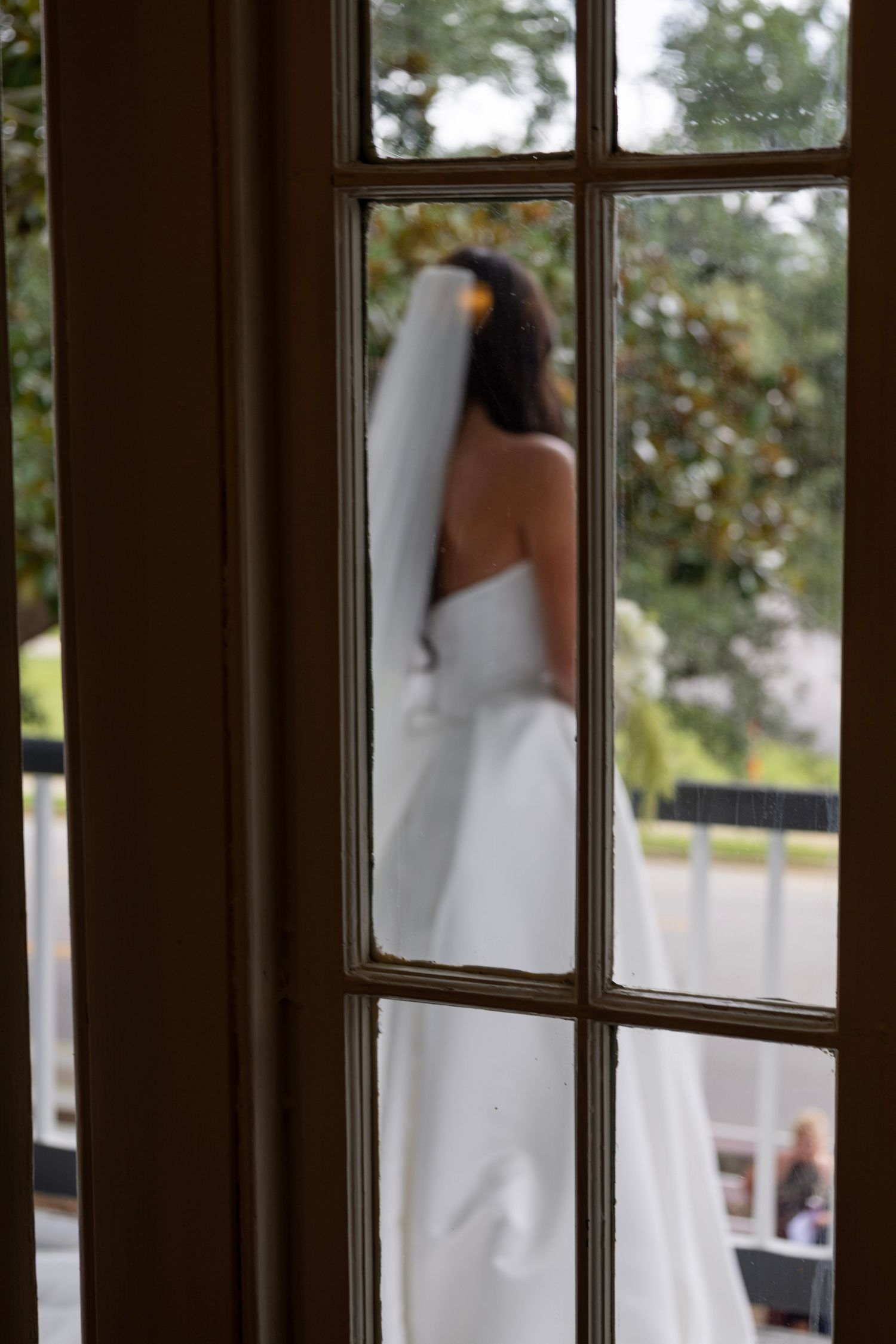 Bride in white wedding dress and veil seen through French doors at outdoor venue.