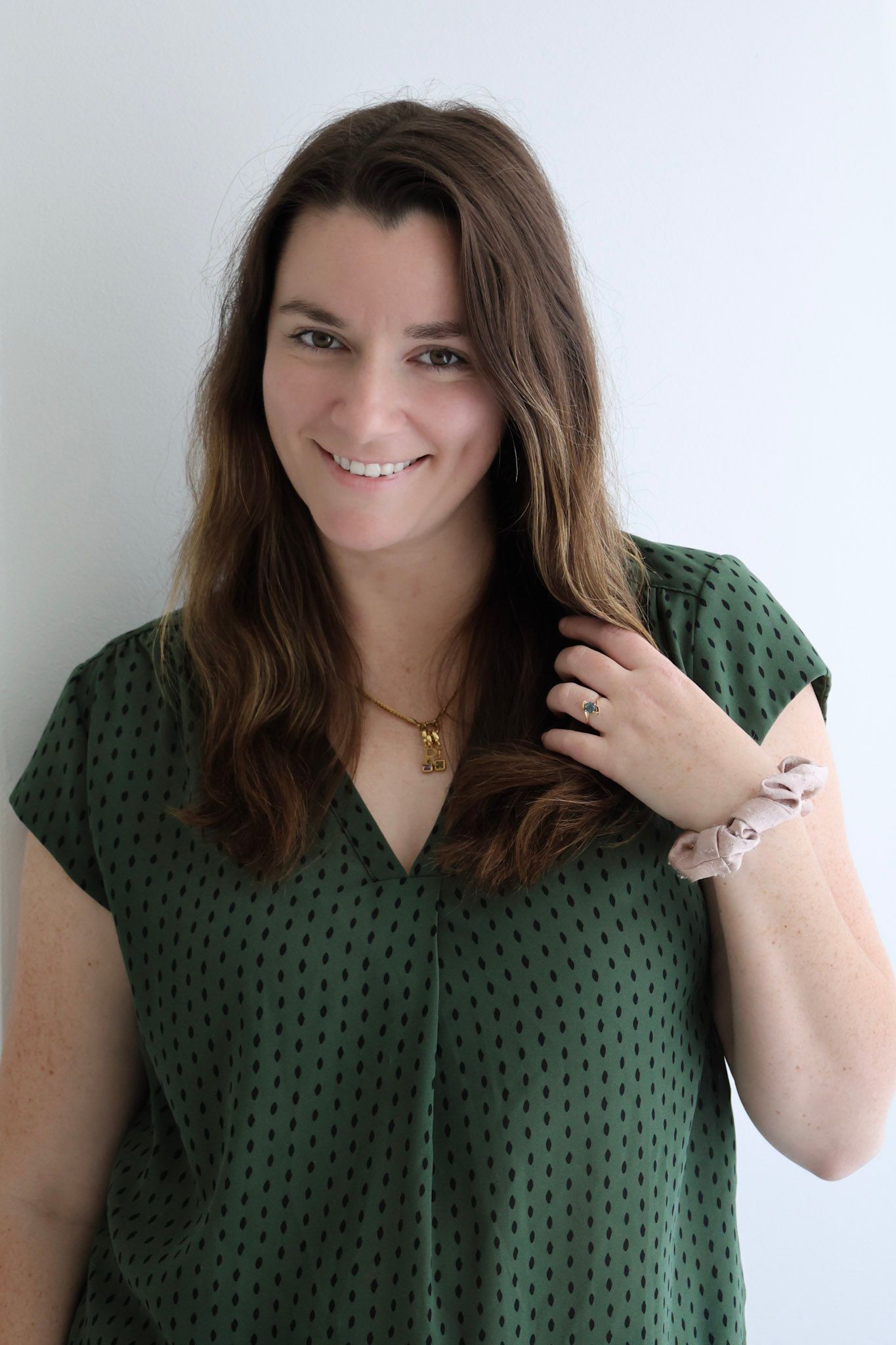Smiling brunette woman in green dotted blouse with gold necklace, touching her hair against white background.