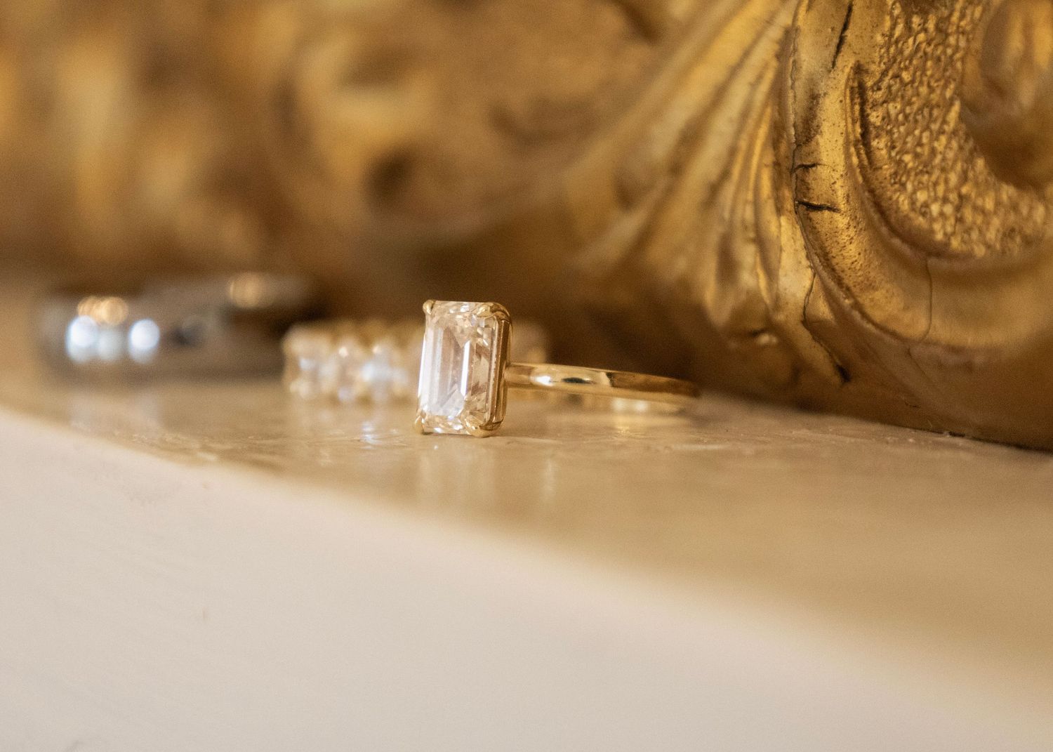 Gold emerald-cut solitaire engagement ring displayed beside a diamond band on a marble surface with ornate gold backdrop.