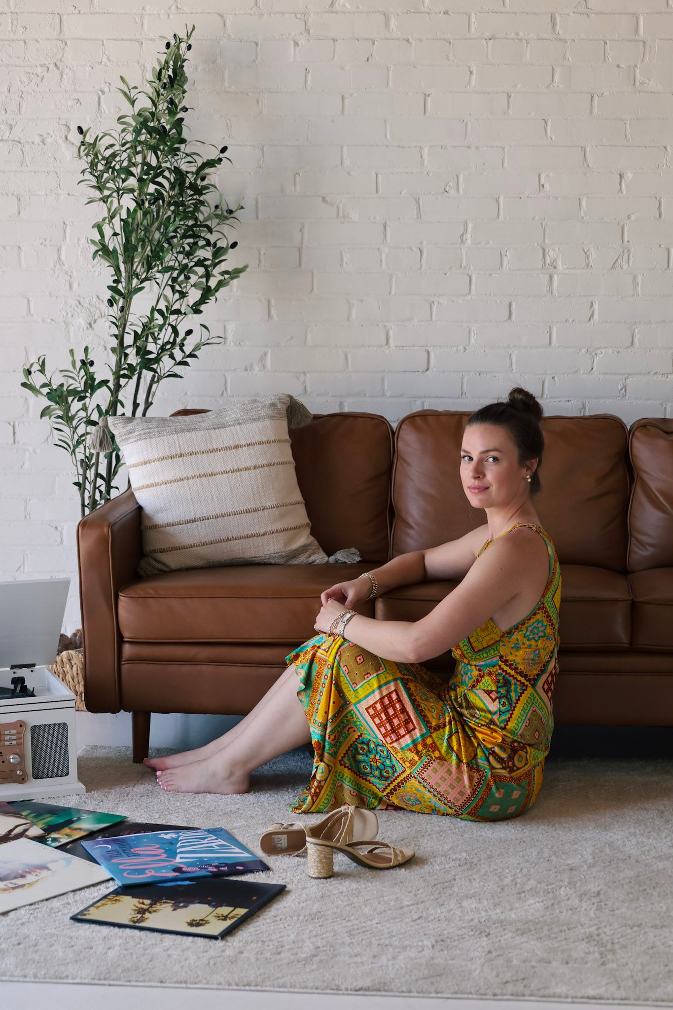 Woman in colorful patterned dress sitting on white carpet beside brown leather sofa with plant and magazines nearby.