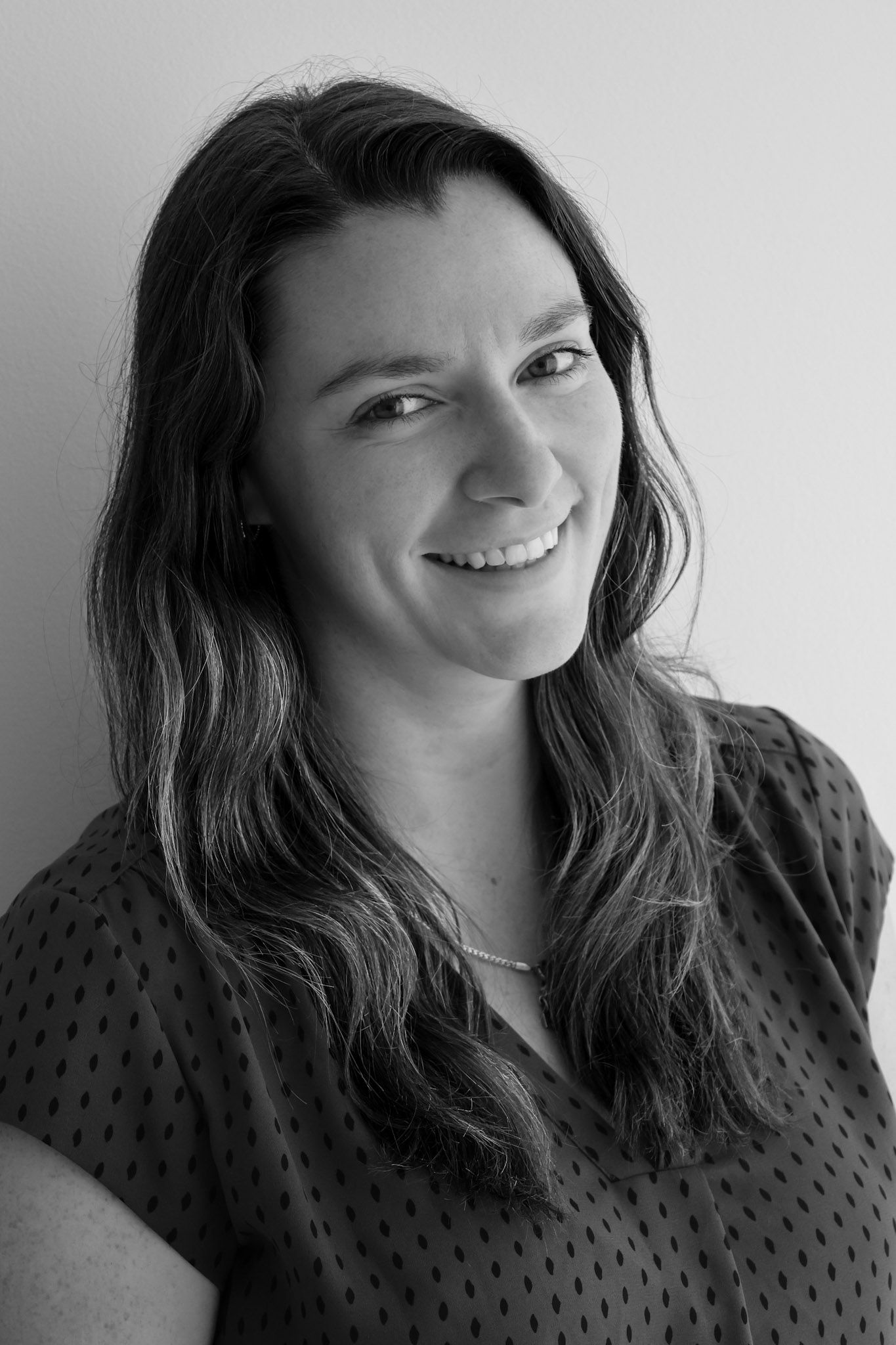 Black and white portrait of a smiling young woman with long hair wearing a polka dot blouse and delicate necklace.