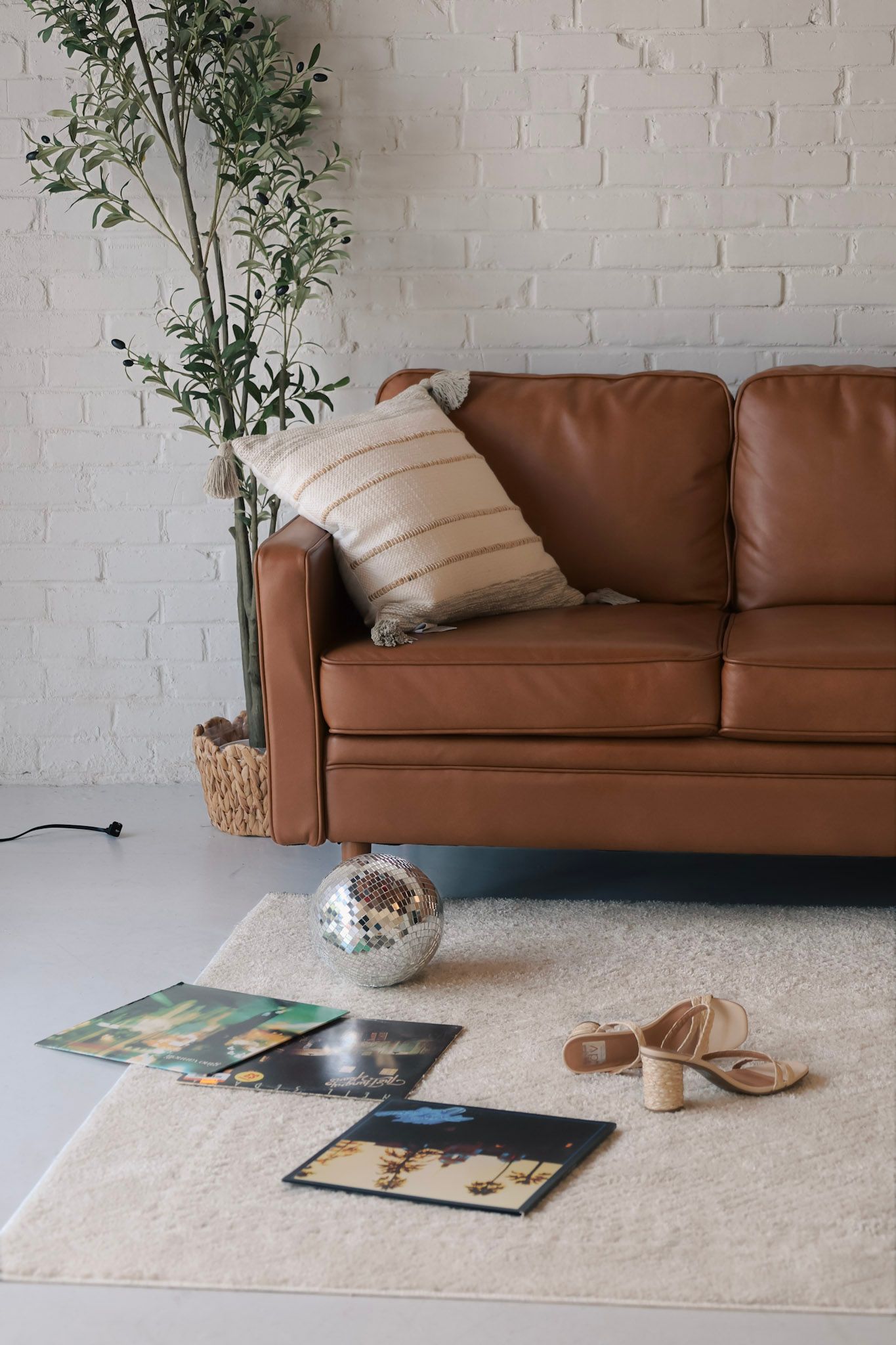 Brown leather sofa with striped pillow, disco ball, vinyl records, and heels on a white shag rug in a modern living room.