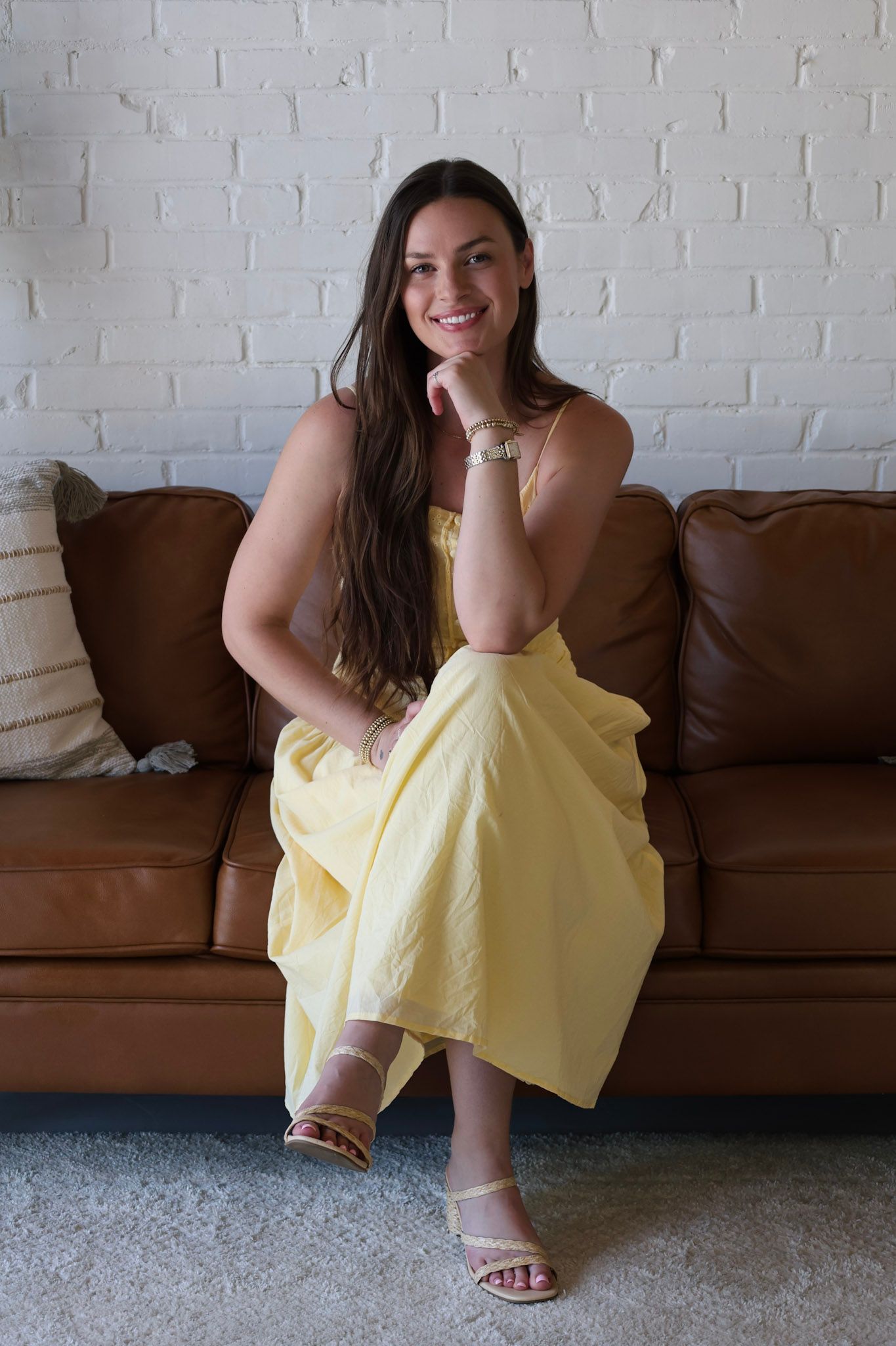 Woman in yellow dress sitting on brown leather sofa, smiling with hand on chin, white brick wall background.