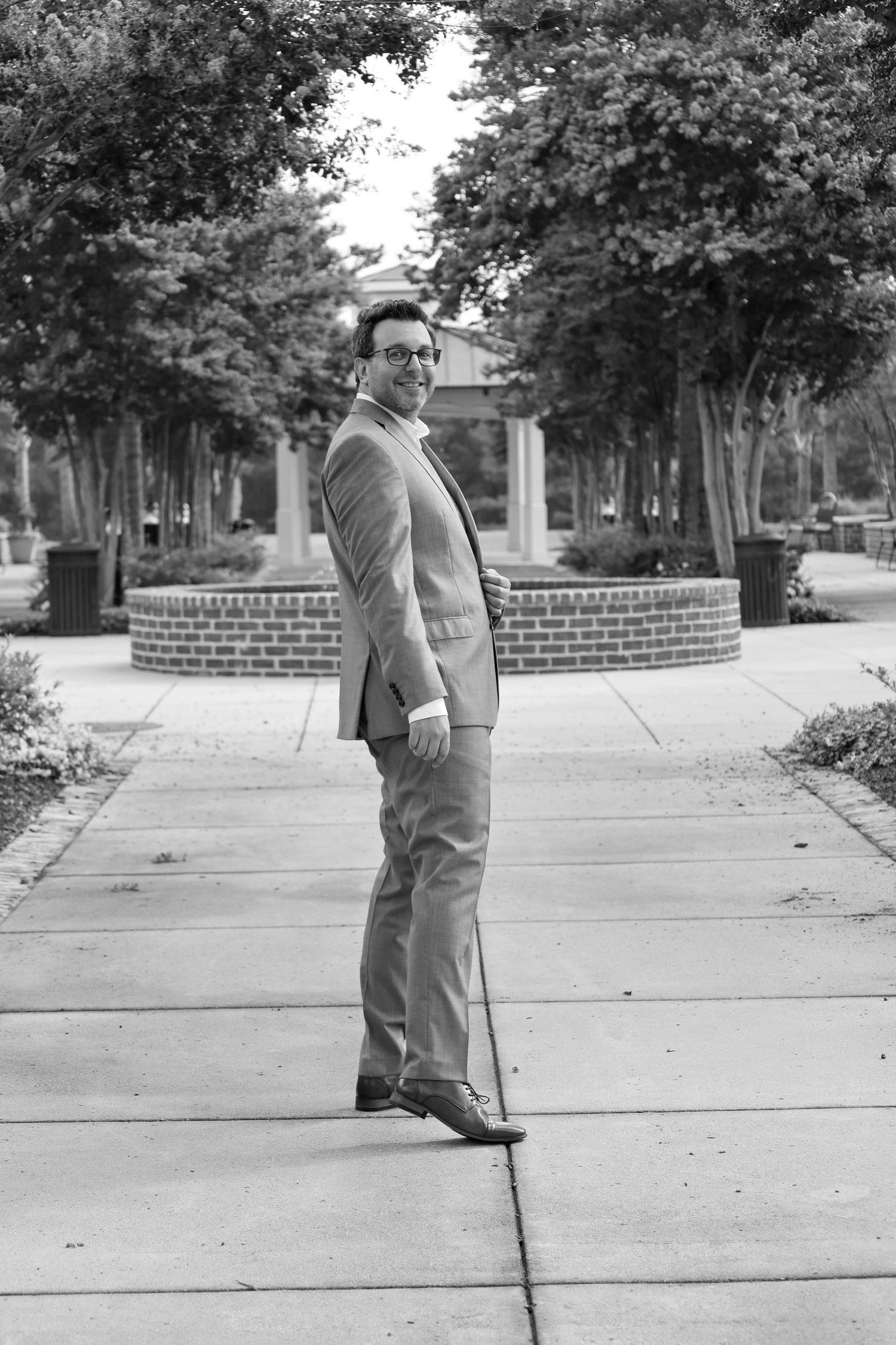 Groom in suit and glasses poses on sidewalk in black and white photo with trees and brick fountain behind him.