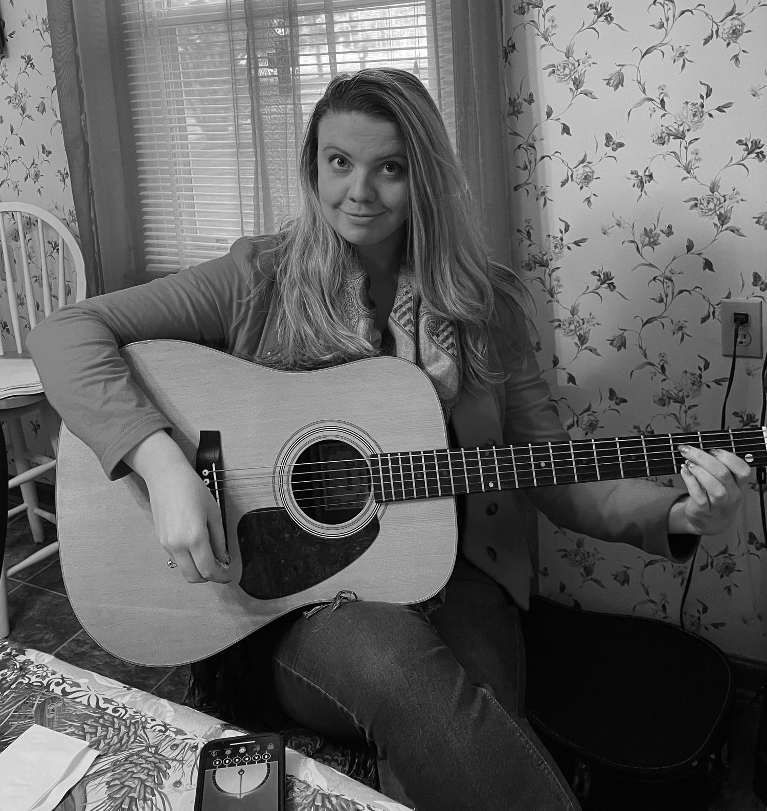 Black and white photo of a young woman smiling while playing an acoustic guitar indoors near a window.