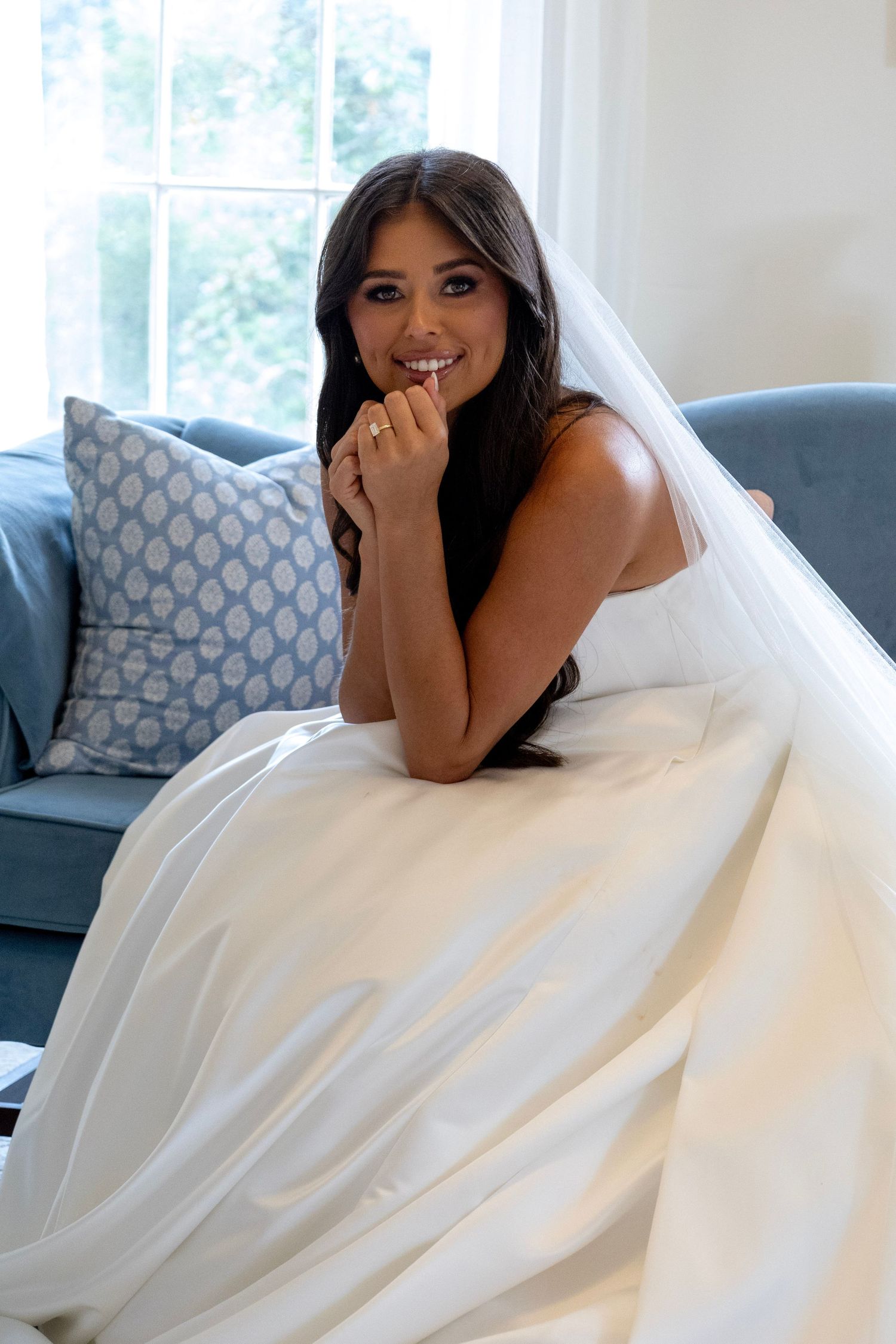 A radiant bride in a white gown and veil sits on a blue sofa, smiling warmly with hands clasped near her face.