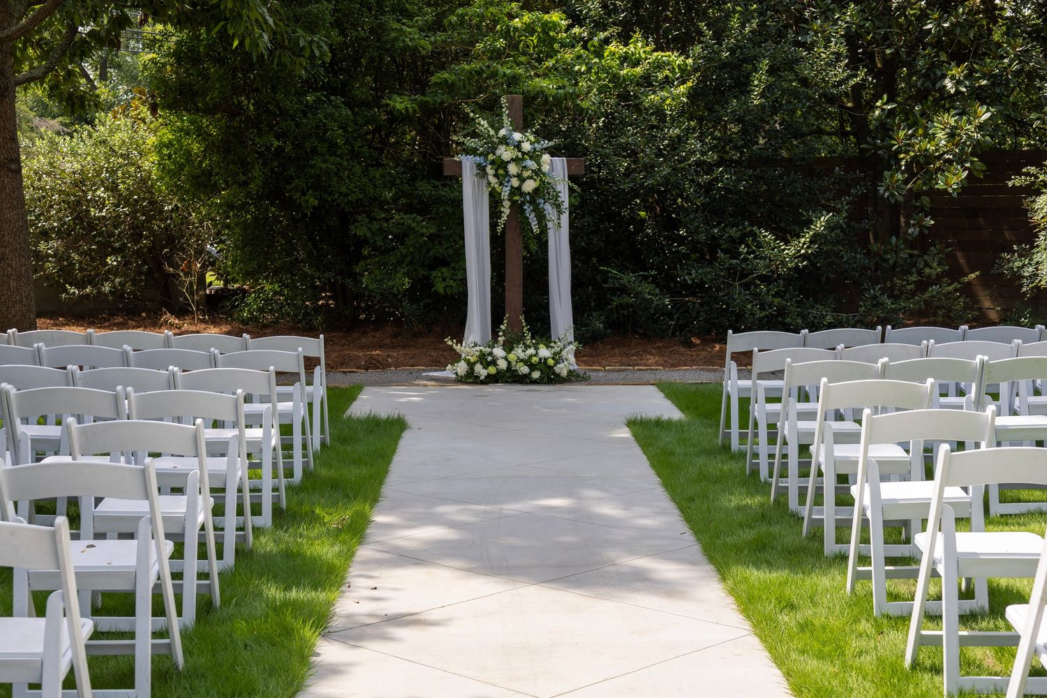 Outdoor wedding ceremony setup with white chairs, a floral arch, and a tree-lined backdrop on a sunny day.
