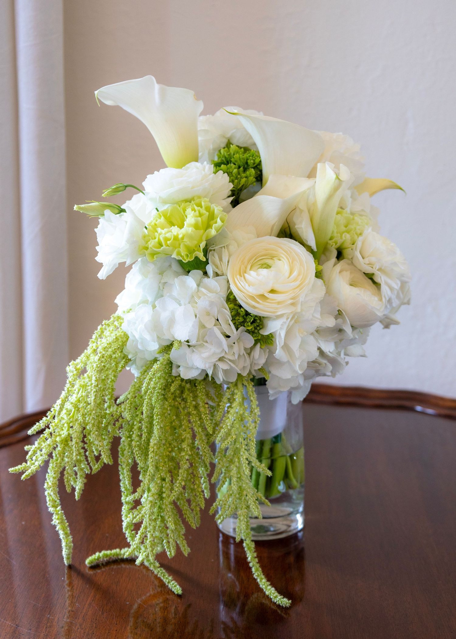 Elegant white and green floral arrangement with calla lilies, roses, hydrangeas, and hanging amaranthus in a glass vase.