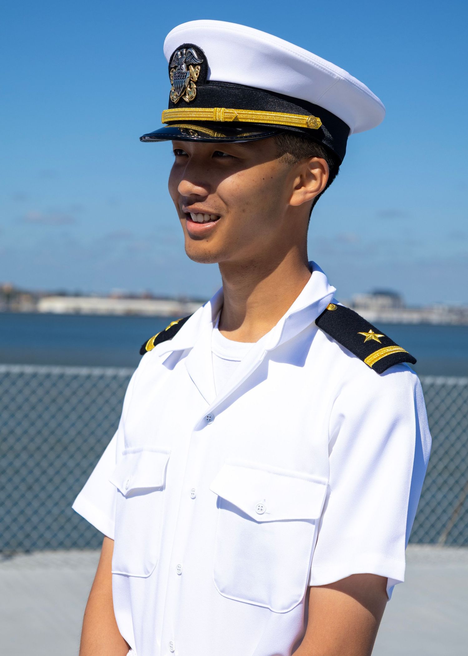 Young naval officer in white dress uniform and cap smiling outdoors near waterfront on a sunny day.