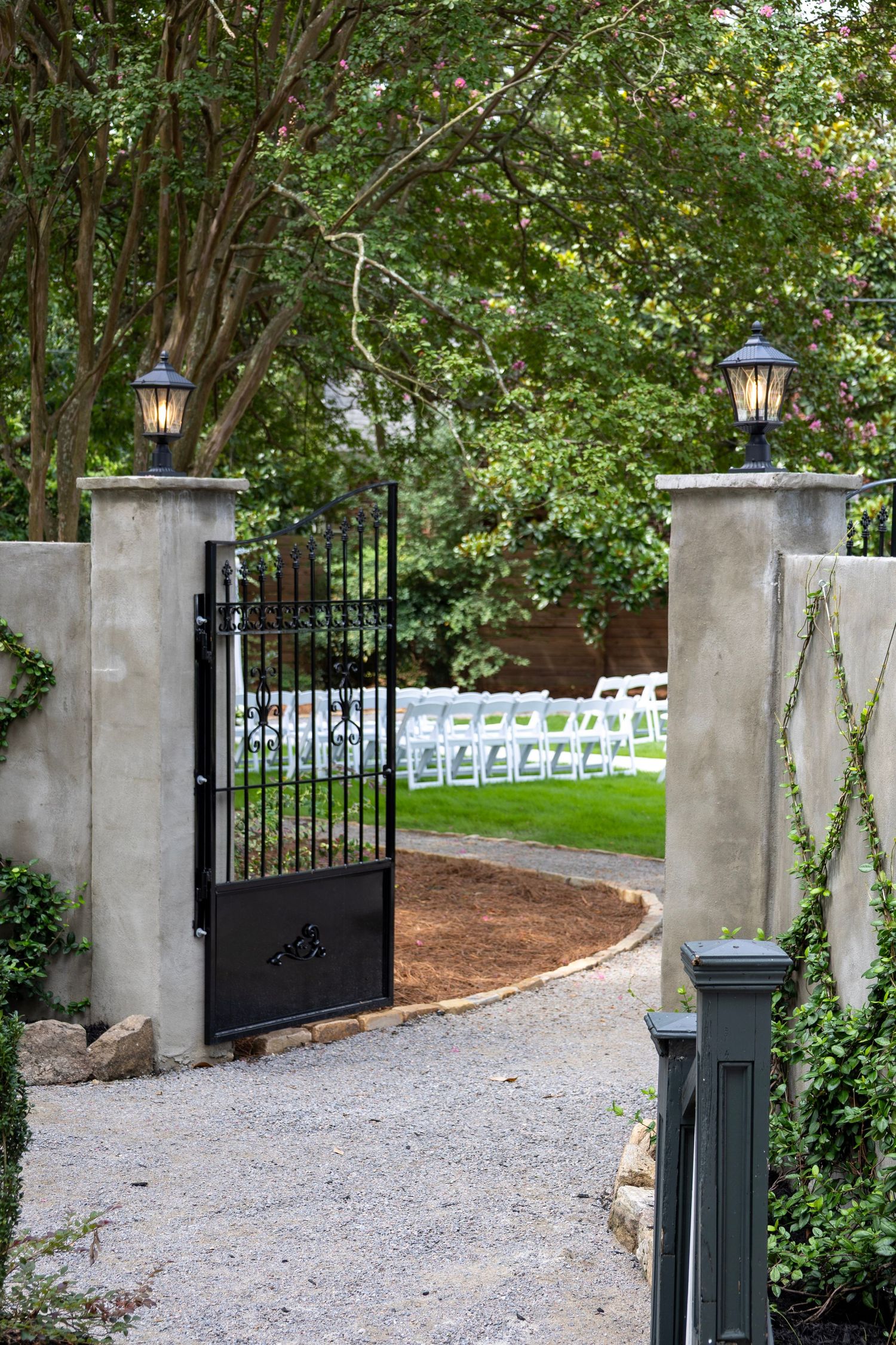 Elegant black iron garden gate with stone pillars and lanterns opening to rows of white chairs for an outdoor wedding ceremony.