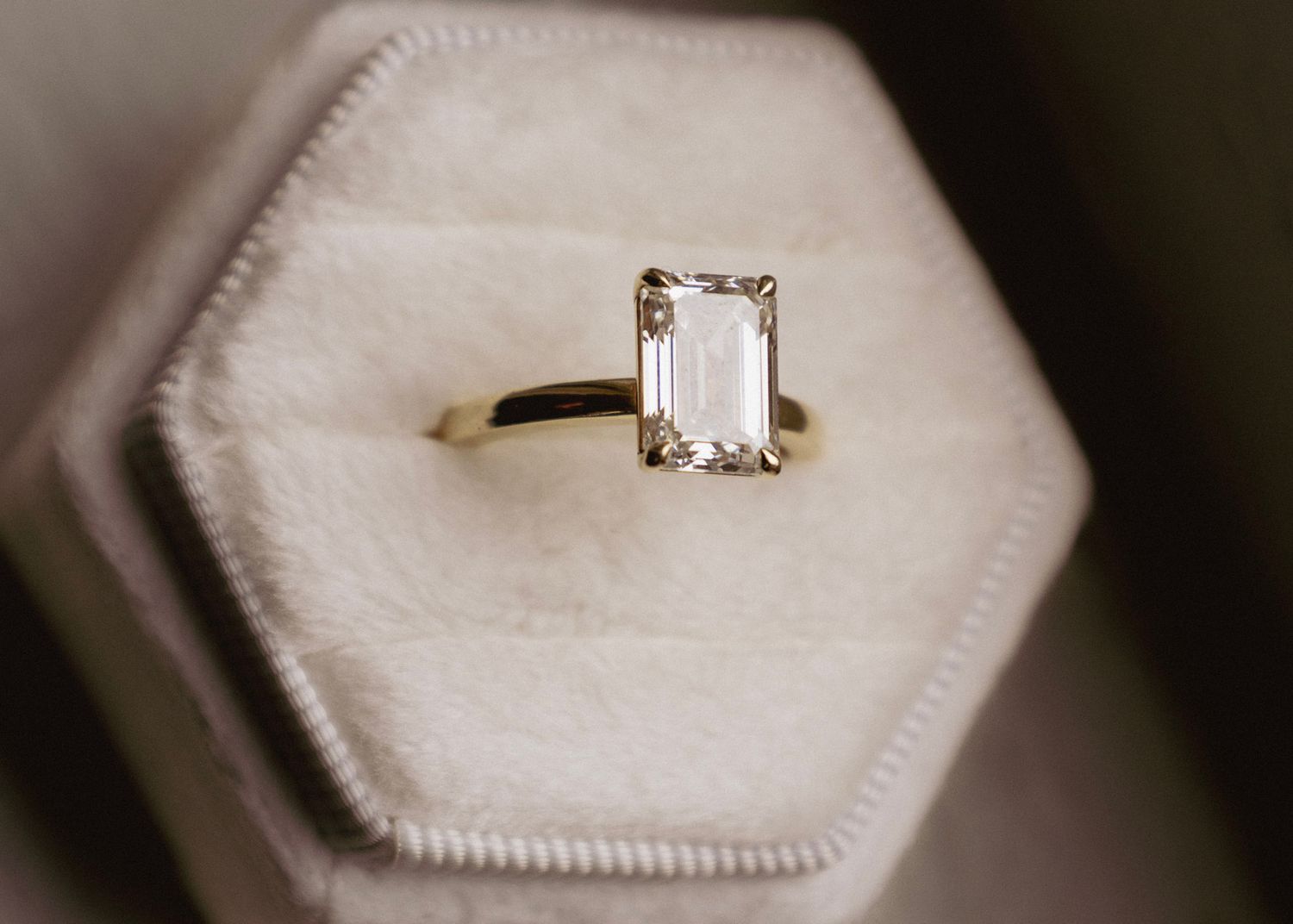 Gold solitaire engagement ring with emerald-cut diamond displayed in a hexagonal velvet ring box.