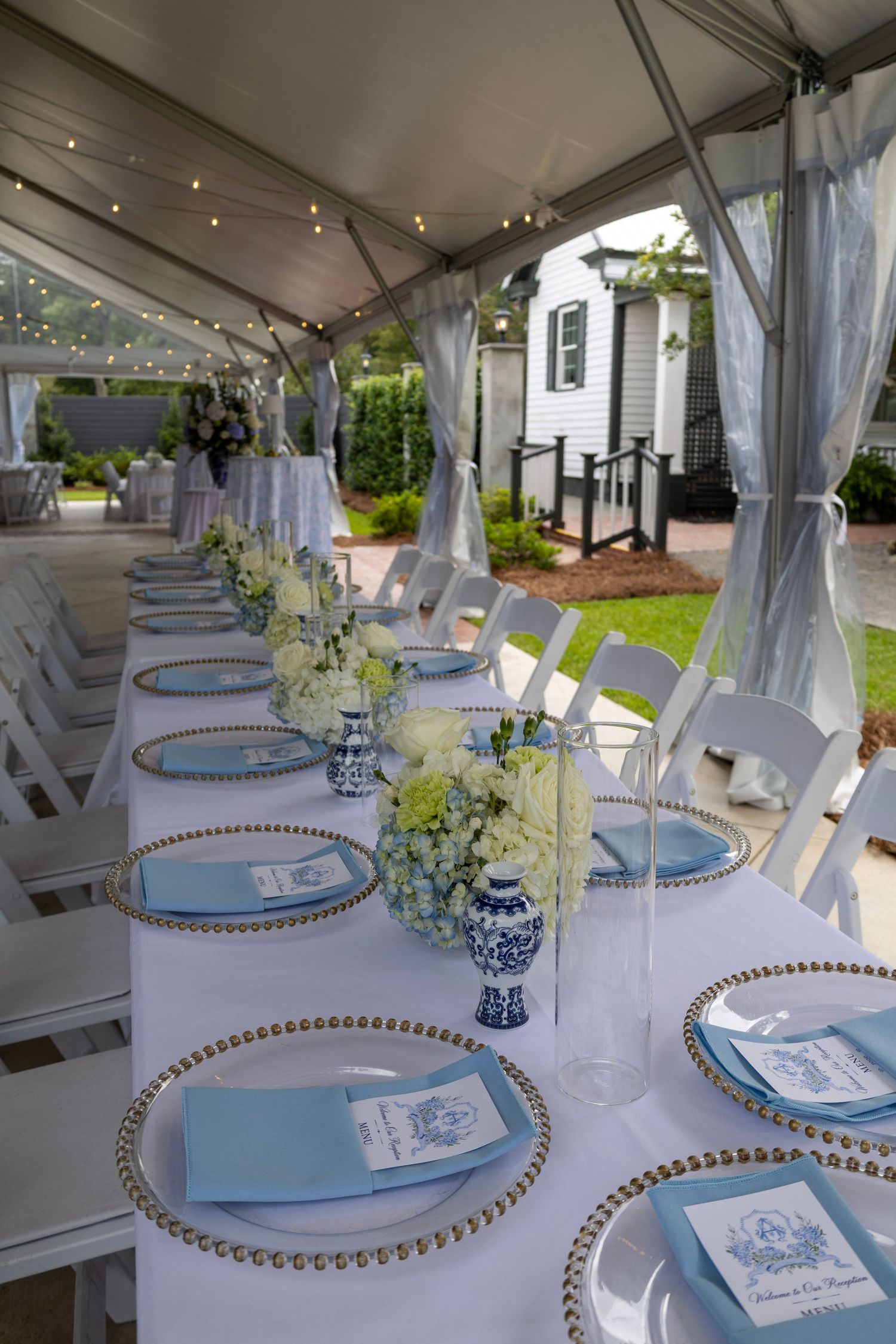 Elegant outdoor wedding reception table with blue napkins, gold-rimmed chargers, and blue-white floral centerpieces under a tent.