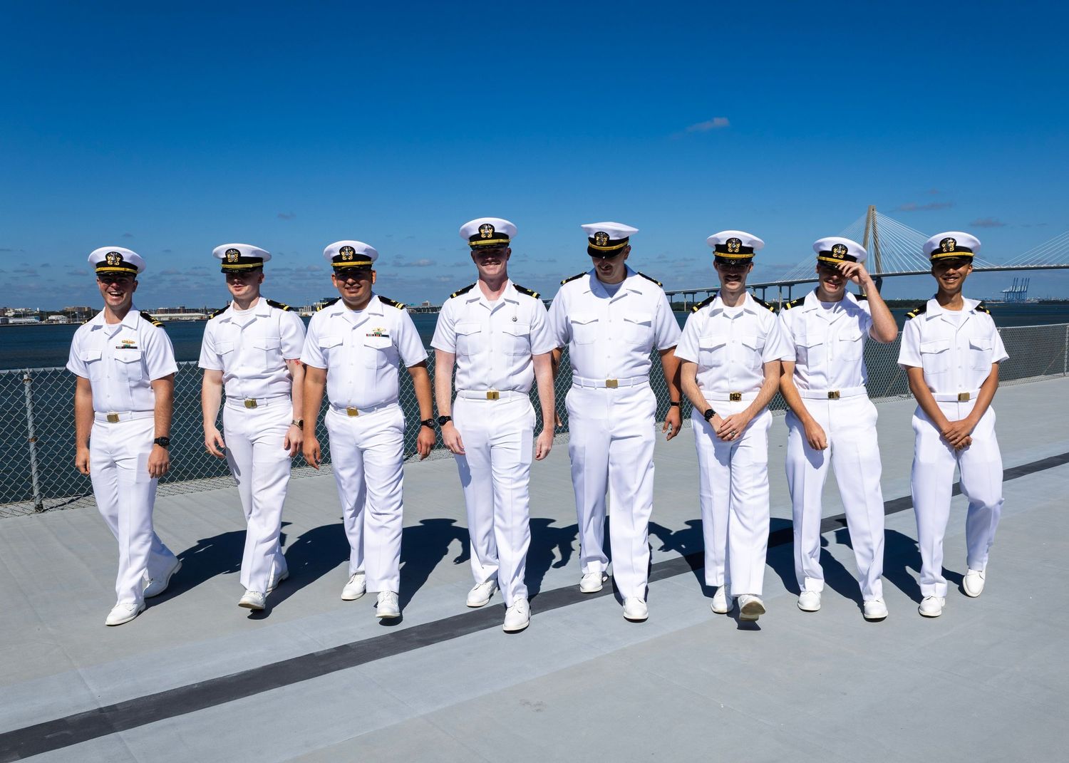 Group of eight Navy officers in white dress uniforms posing on a ship deck with a bridge visible in the background.