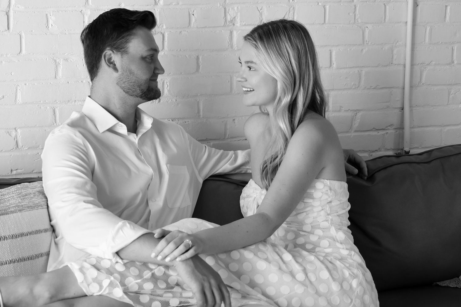 Black and white photo of a couple sitting together on a couch, smiling and holding hands romantically.