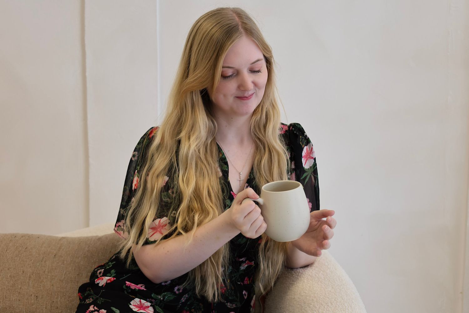 Young blonde woman in floral dress smiling while holding a ceramic mug, seated on a cozy chair.
